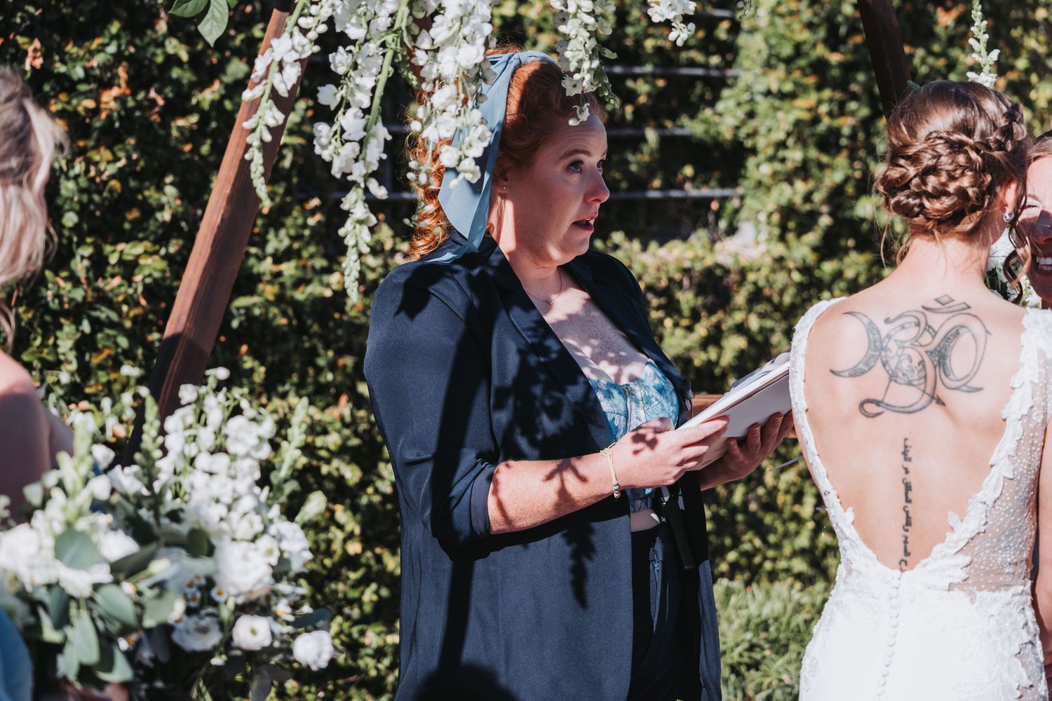 Tattooed officiant in black cape reads vows from book beside bride in lace backless gown at outdoor wedding.