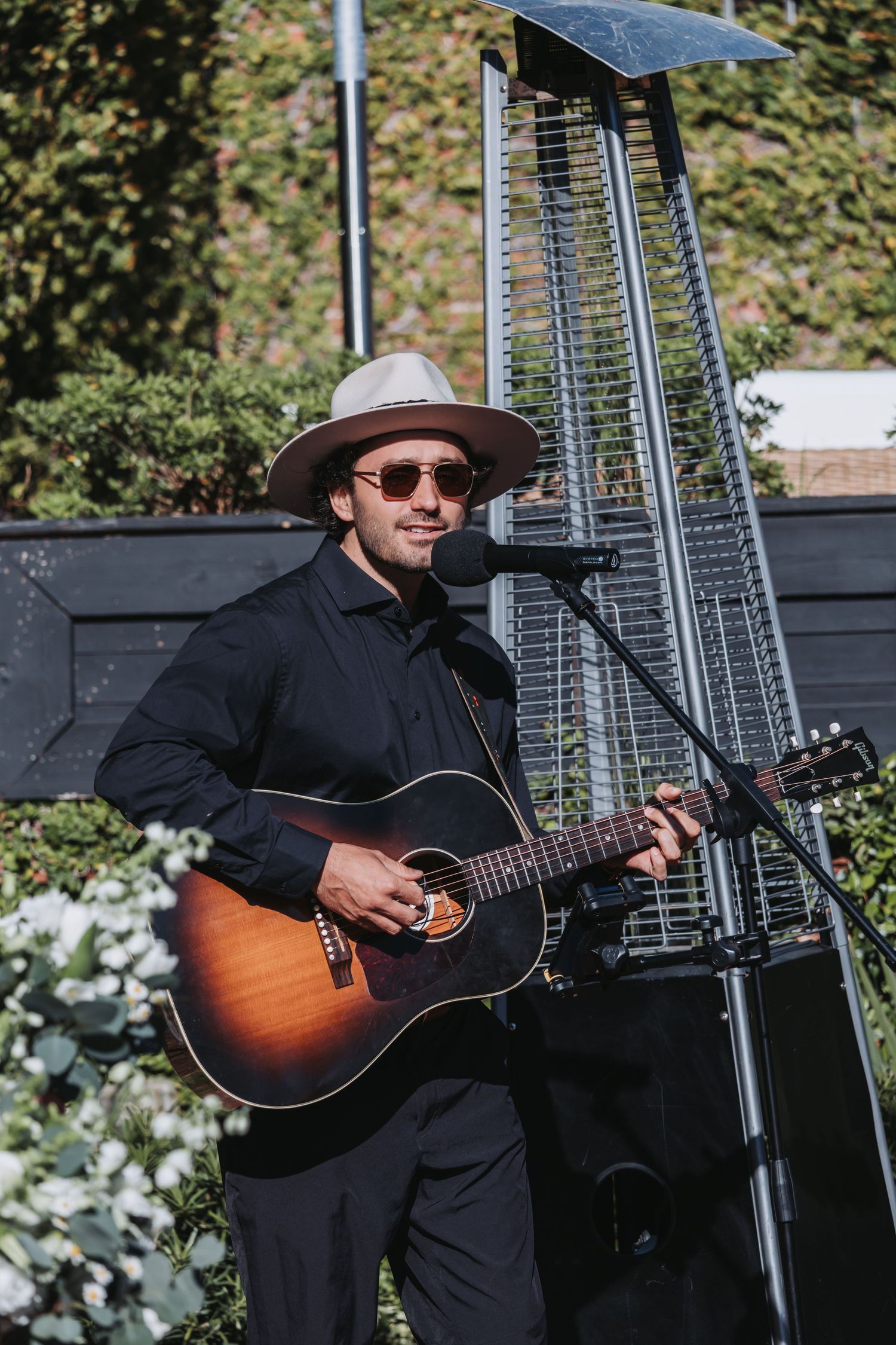 Man in hat and sunglasses playing acoustic guitar at outdoor microphone stand near patio heater.