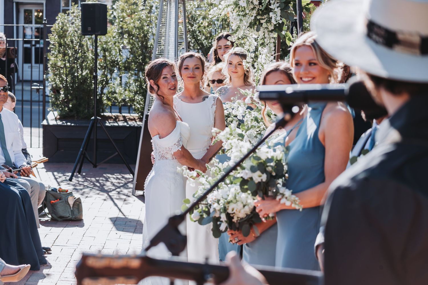 Bride in white gown stands with bridesmaids holding floral bouquets during an outdoor wedding ceremony.