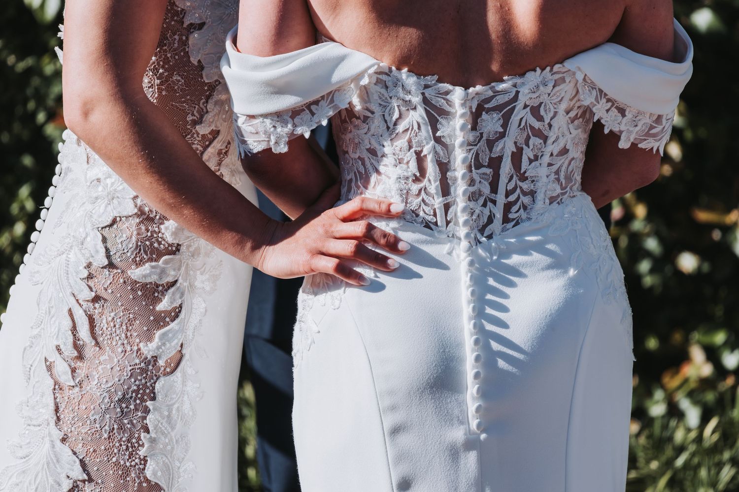 Close-up of two brides in white lace wedding dresses embracing during their wedding ceremony outdoors.