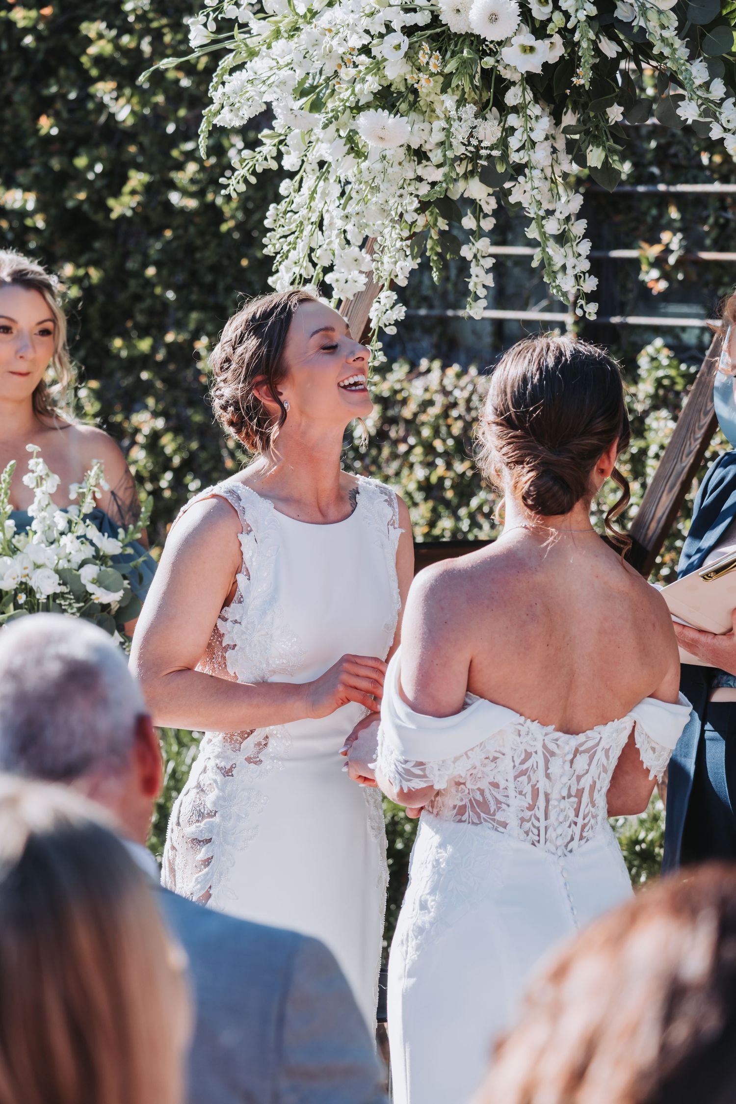 Two brides in white wedding gowns exchange vows under a floral arch at an outdoor wedding ceremony.