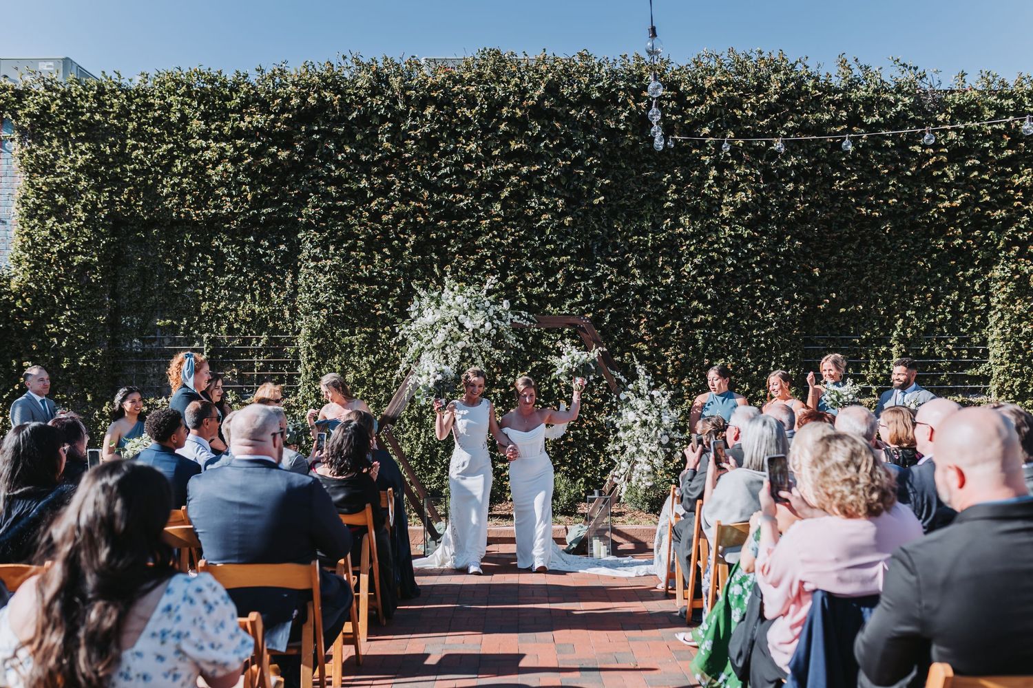 Two brides walk back down the aisle after their wedding ceremony in a garden venue lined with guests.