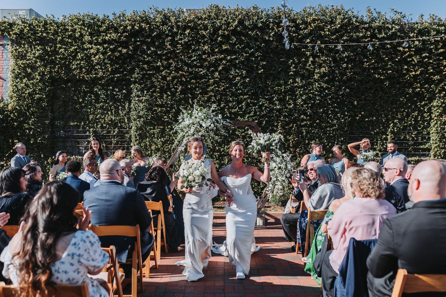 Two brides in white gowns walk hand in hand down brick aisle after wedding ceremony before ivy-covered wall.