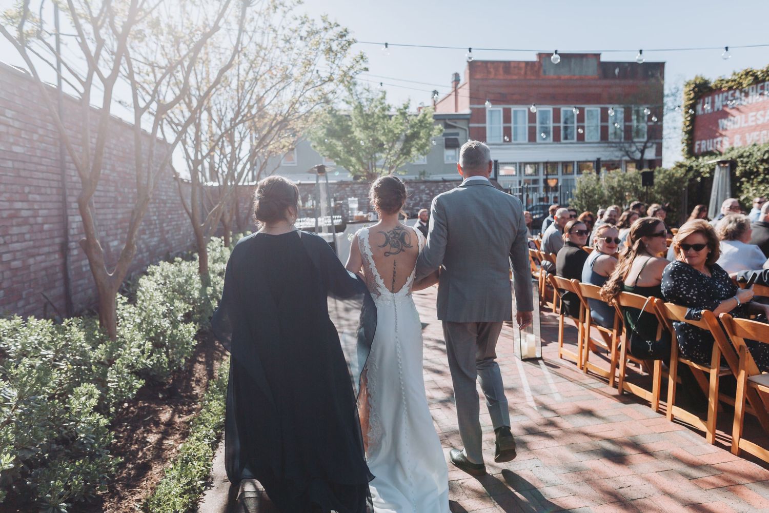 Bride in elegant backless gown walks down outdoor aisle escorted by two guests at a brick venue wedding.