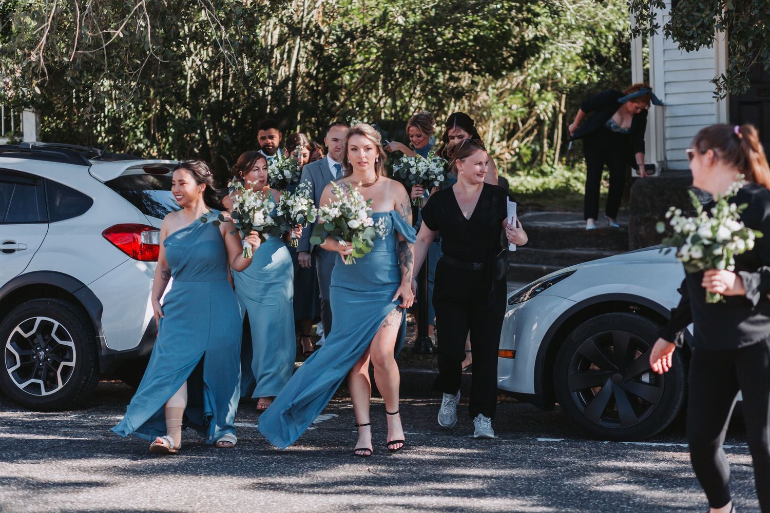 Bridesmaids in blue dresses carrying white bouquets walk together outdoors near cars at a wedding.