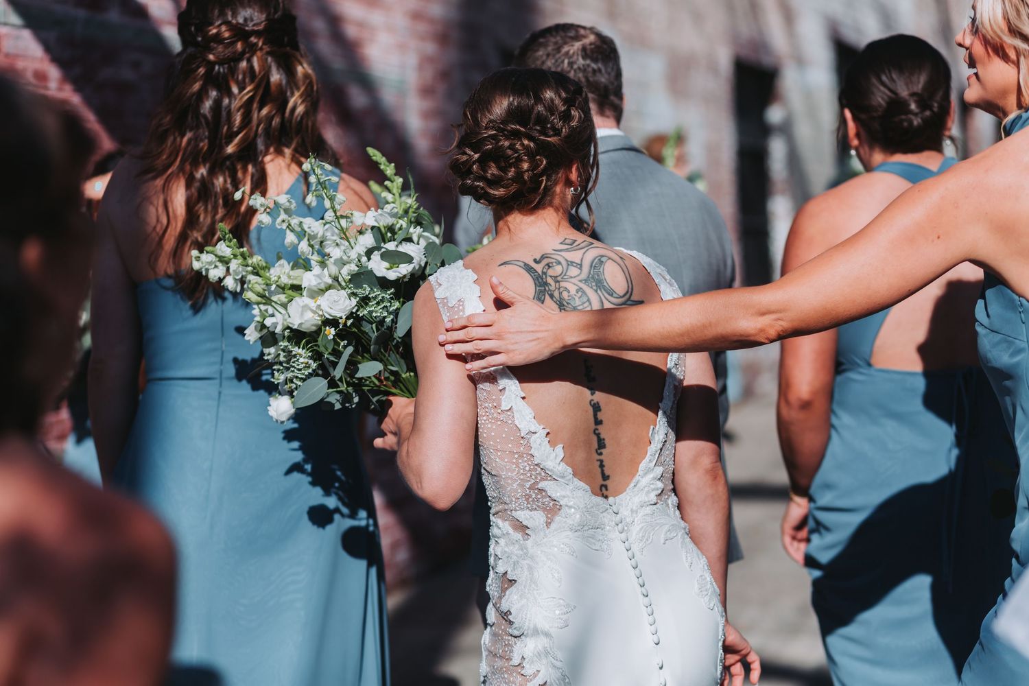 Bride with back tattoo in lace wedding dress walks with bridesmaids in blue dresses holding white floral bouquet.