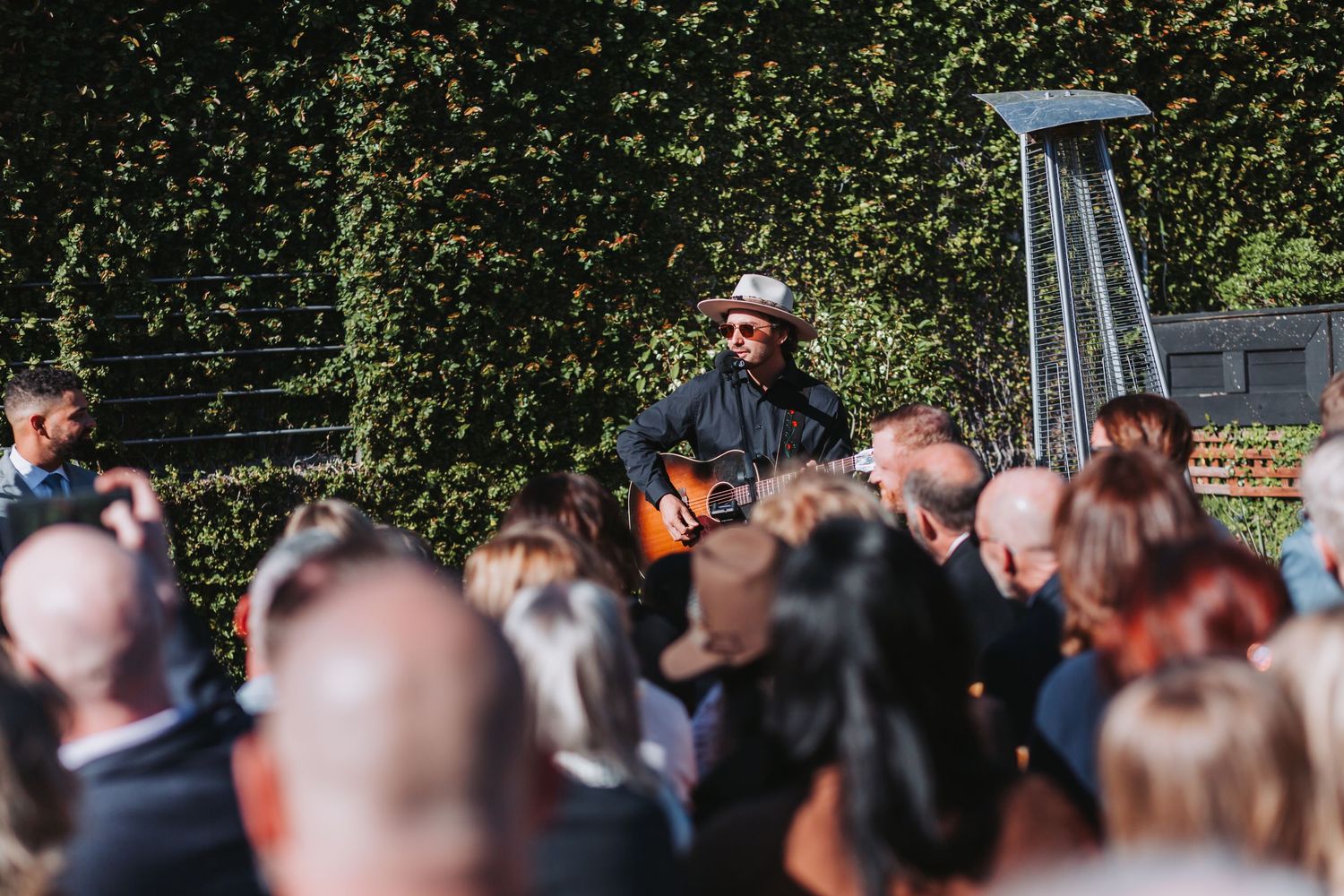 Musician playing acoustic guitar outdoors for a crowd at a sunny garden event.