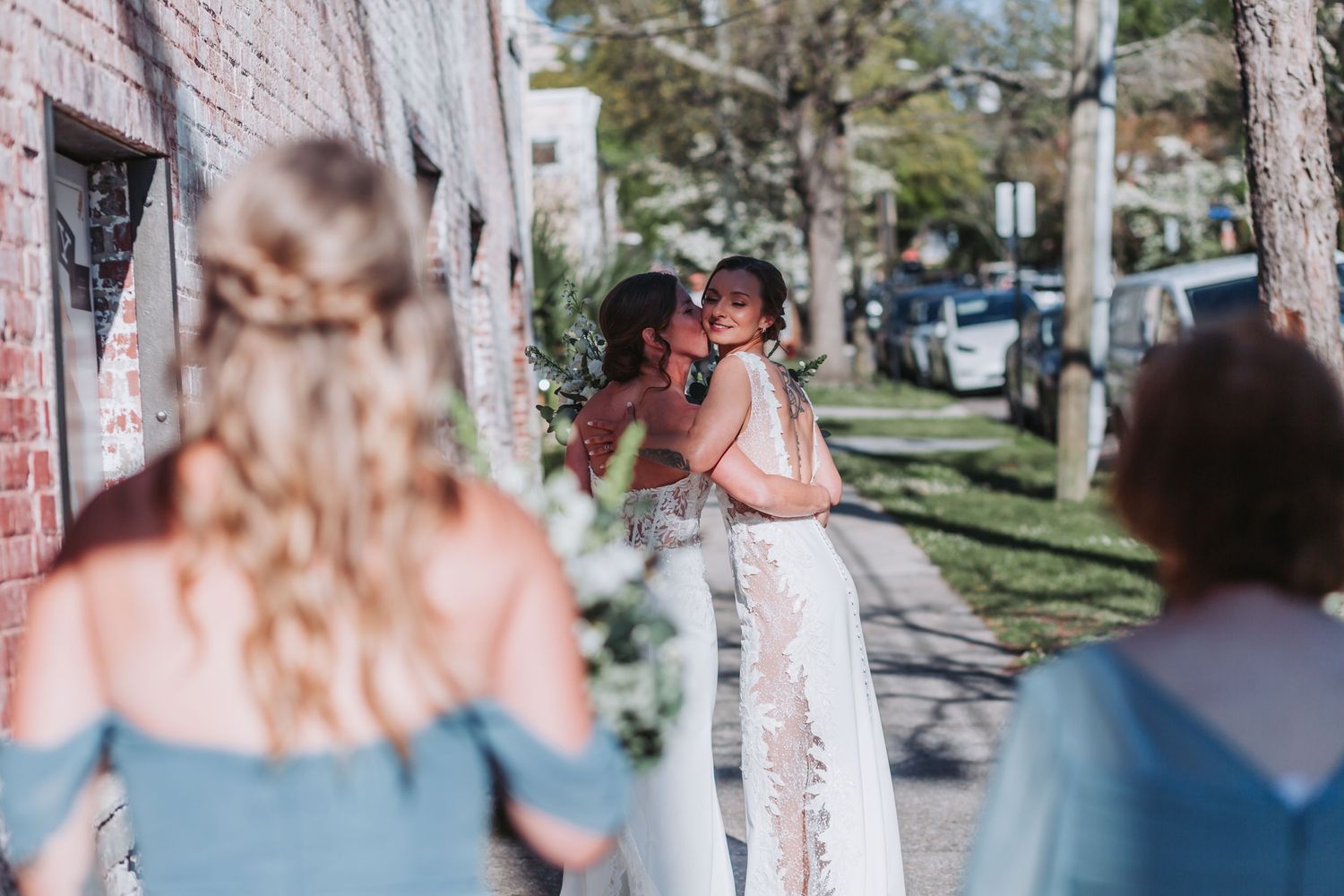 Two brides share a kiss outdoors while bridesmaids in blue dresses look on during a wedding celebration.