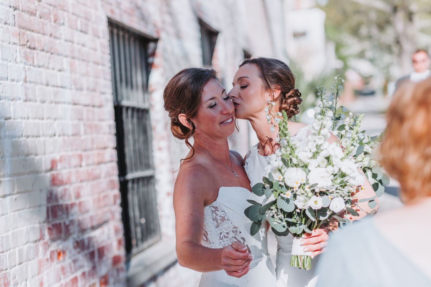 Two brides sharing a tender kiss outdoors, one holding a white and greenery wedding bouquet near a brick wall.
