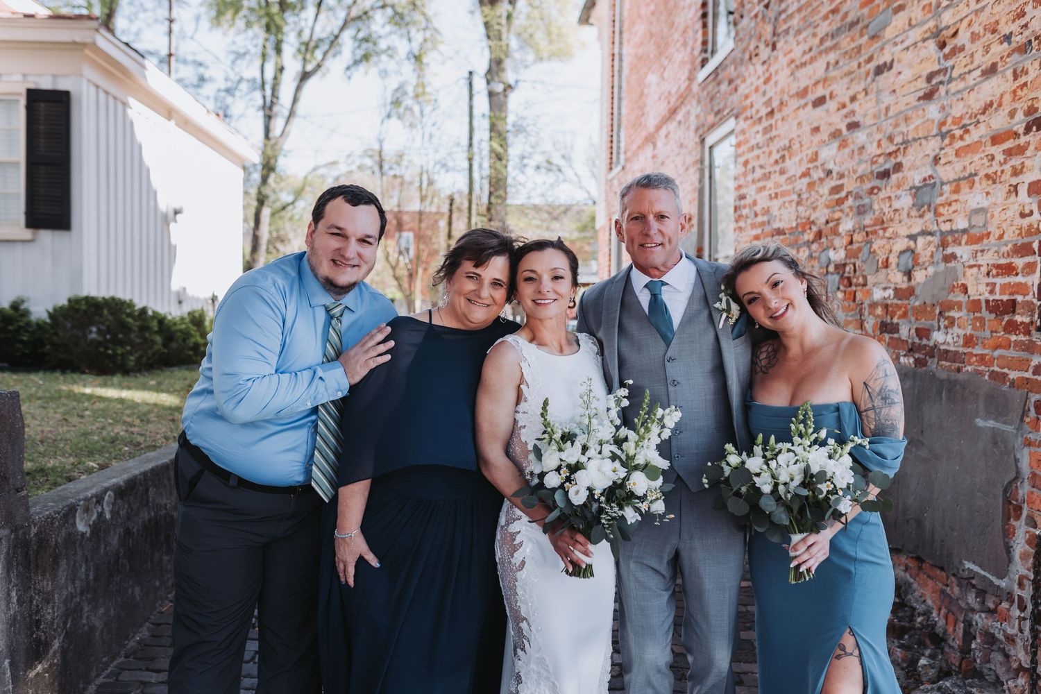 Bride with family in dusty blue attire holding white bouquets outside brick church.