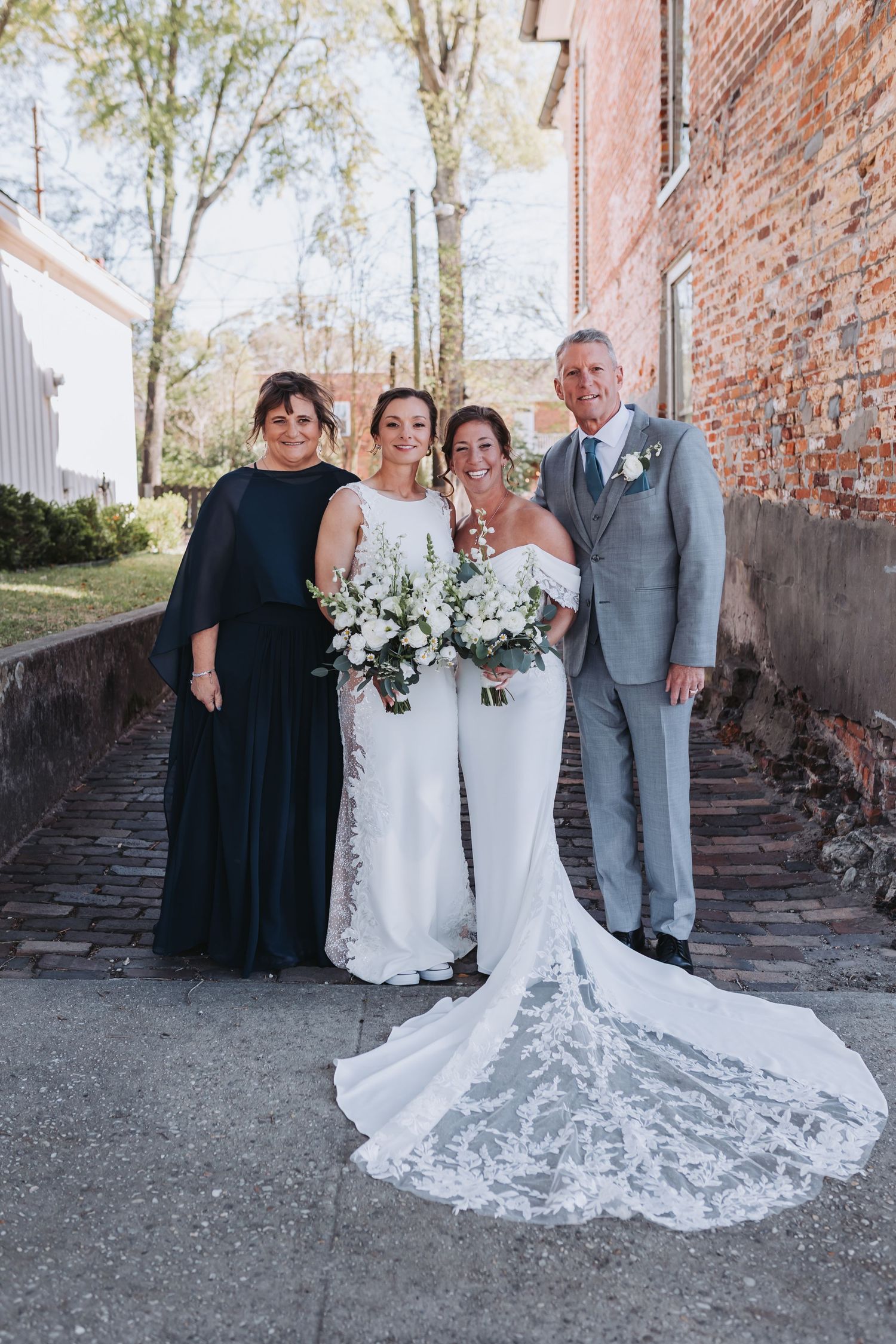 Two brides in white gowns pose with family outside a brick building on their wedding day.