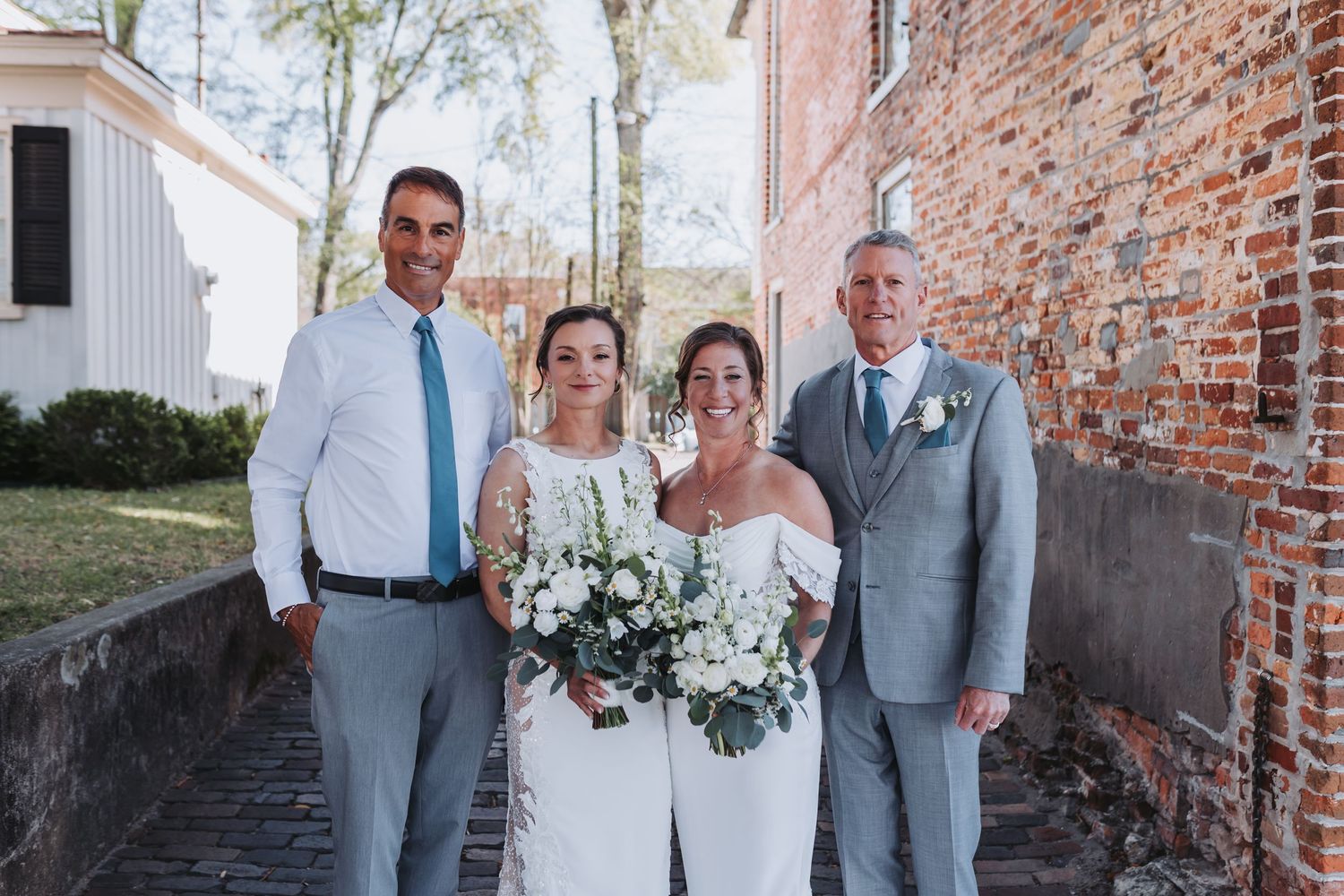Wedding party of four posing outdoors near brick wall, bride holding white floral bouquet in elegant off-shoulder gown.