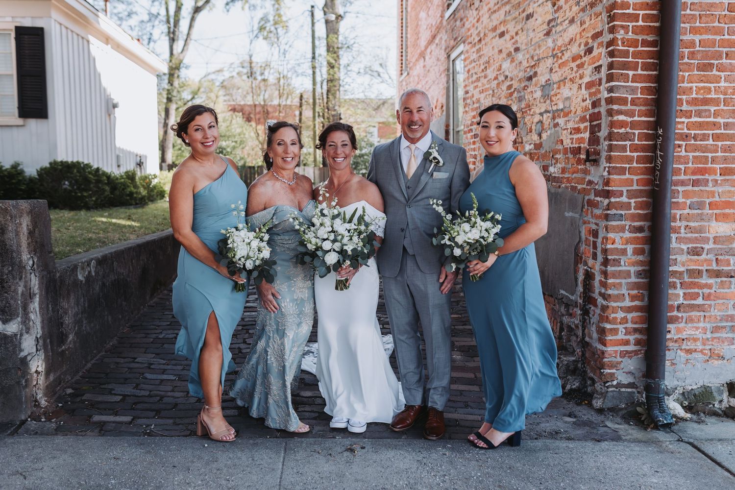 Bride and groom pose with two bridesmaids in blue dresses holding white bouquets outside a brick building.