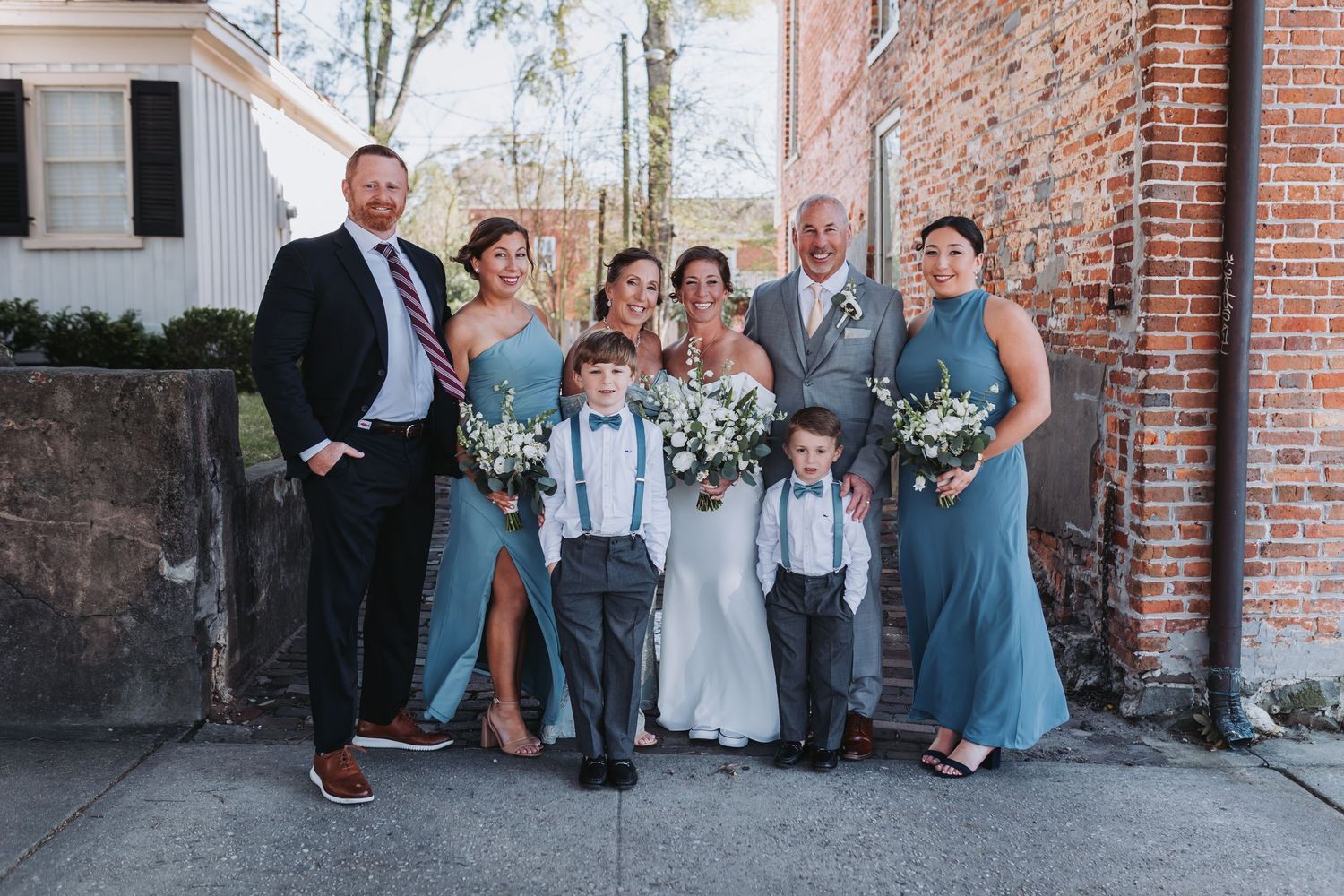 Wedding party poses outdoors in dusty blue gowns and gray suits, with bride in white holding greenery bouquet.
