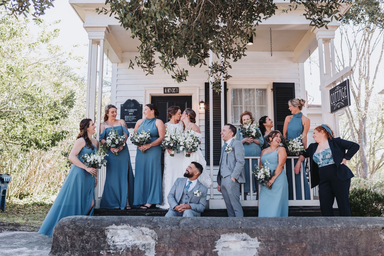 Large bridal party in dusty blue dresses and gray suits posing outside white historic building with bouquets.