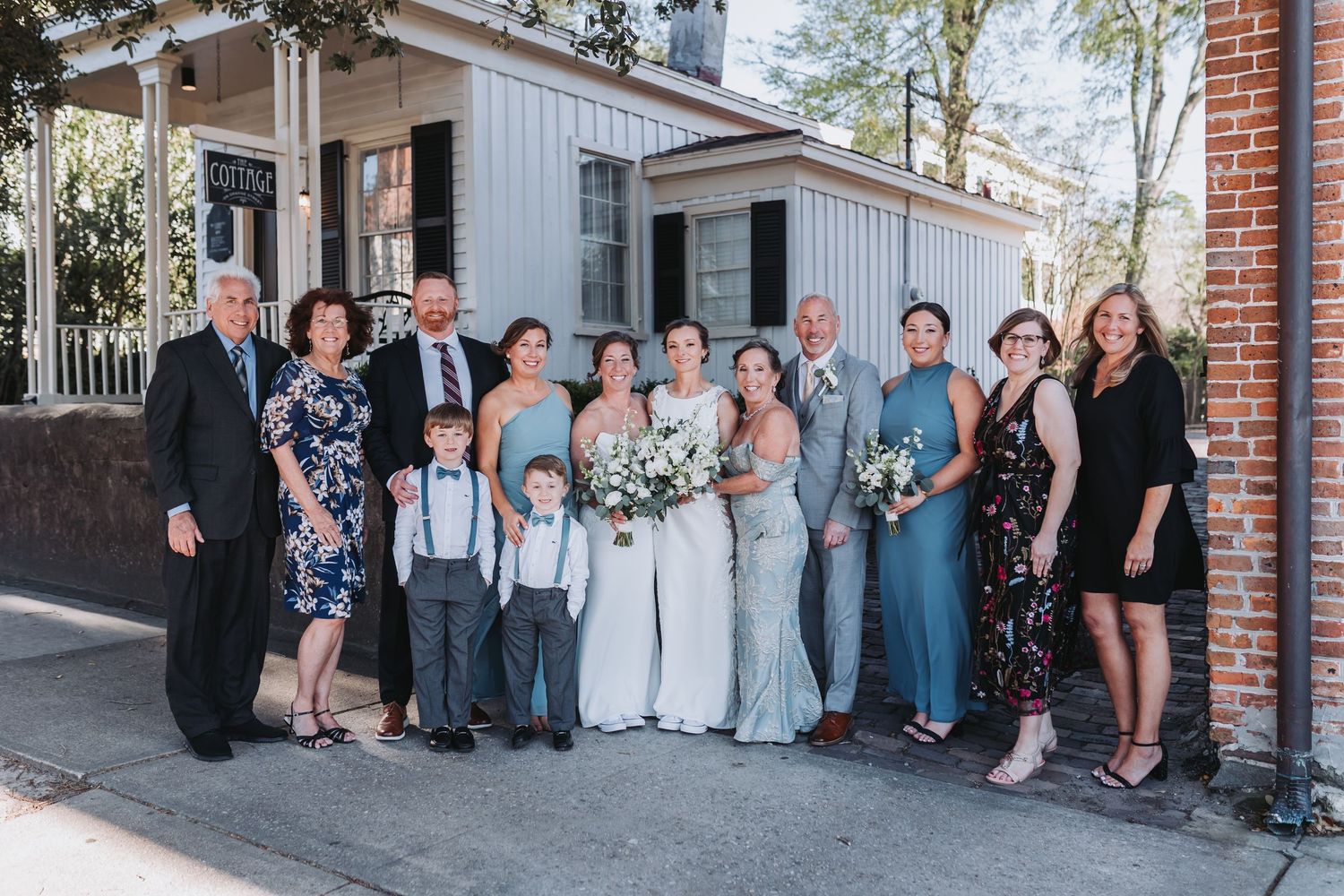 Wedding party group photo outdoors, featuring bride, groom, and family members in formal attire near a brick building.