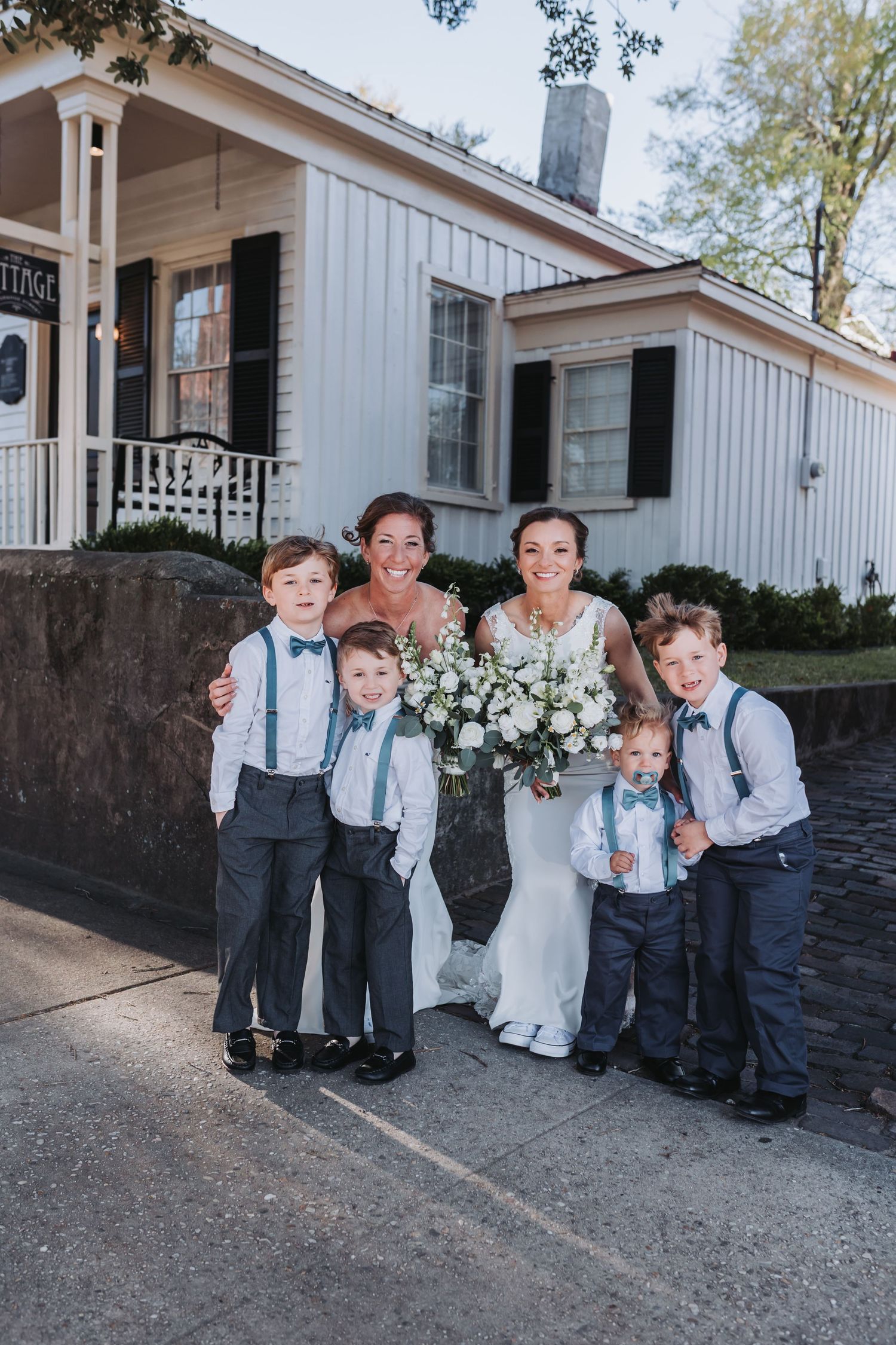Two brides in white gowns pose with four young ring bearers in navy suspenders and bow ties outside a white venue.