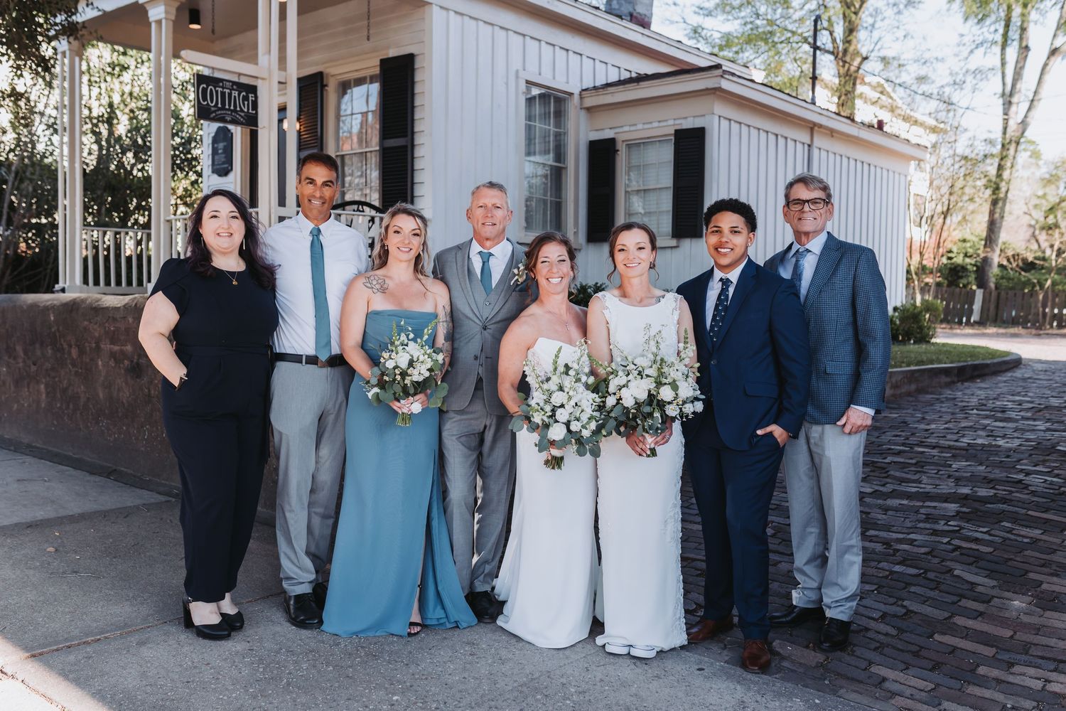 Wedding group portrait of eight people outdoors, two brides holding white bouquets flanked by family members.