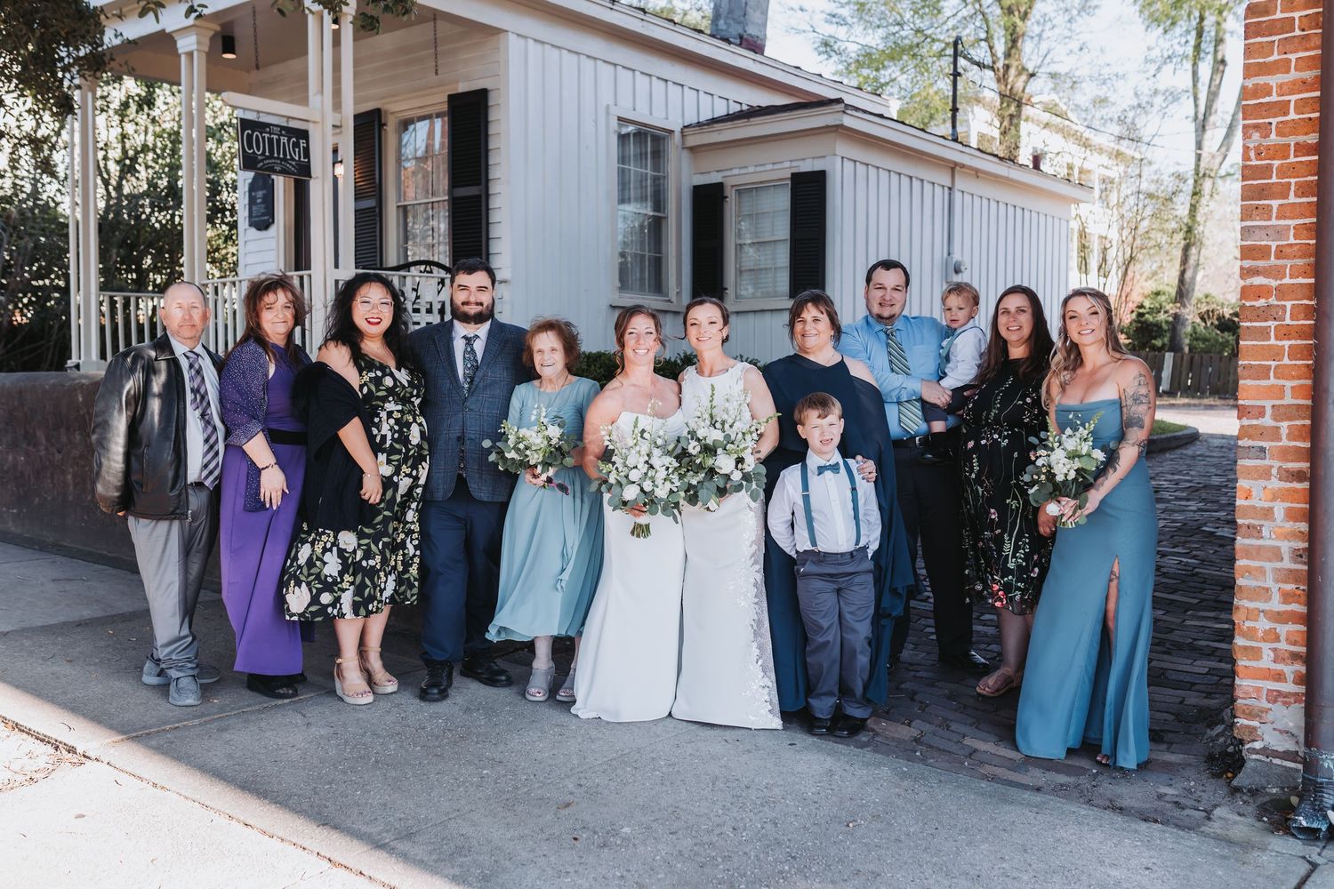 Wedding party poses outside a historic building, bride in white gown holding bouquet, guests in blue and purple attire.