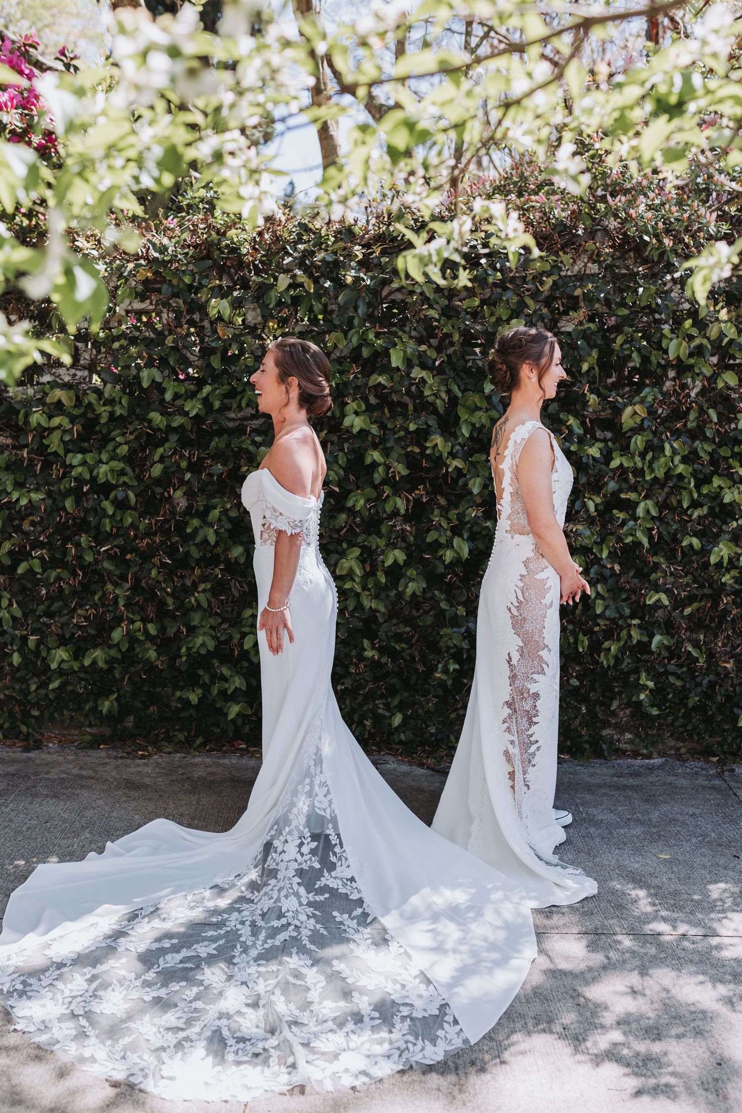Two brides in elegant lace wedding gowns with long trains stand back-to-back before a lush green hedge.