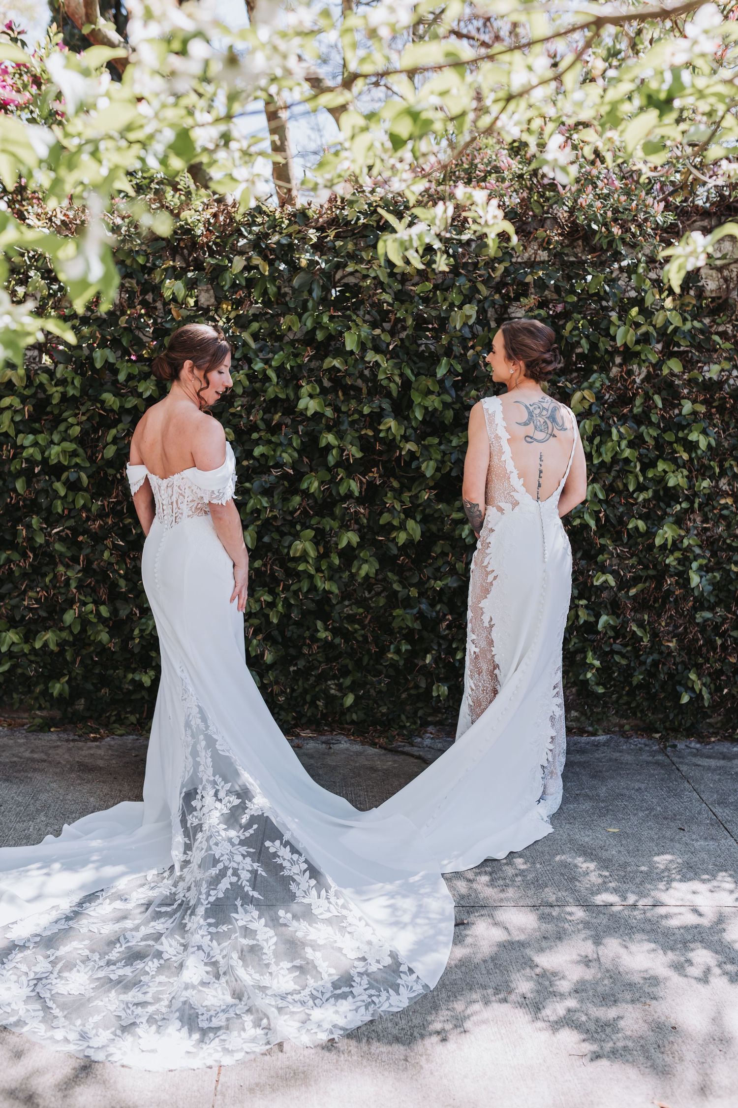 Two brides in elegant white lace wedding gowns with long trains stand back-to-back in a lush garden.