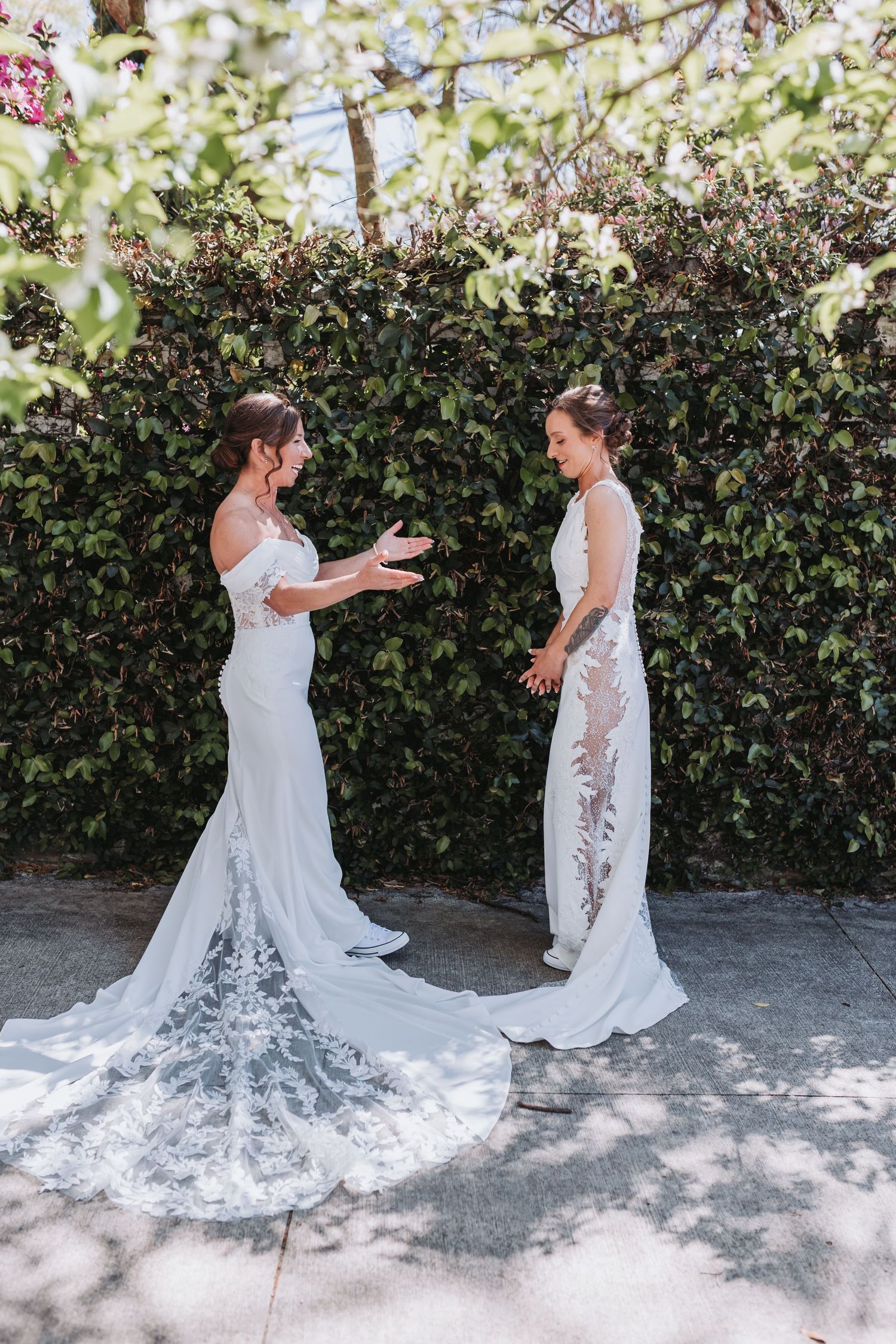Two brides in elegant white wedding gowns share a joyful moment in a lush garden setting.