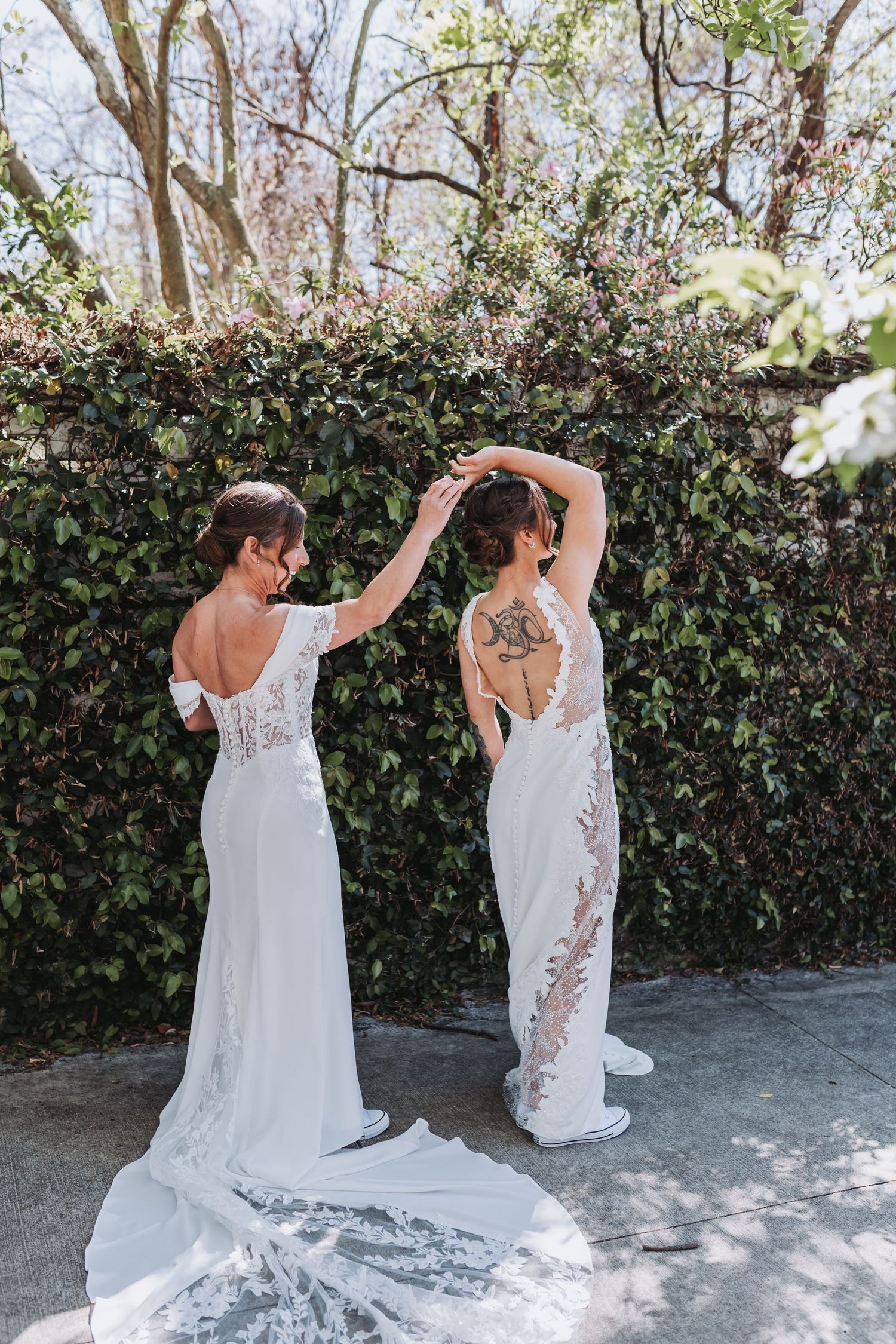 Two brides in white wedding gowns pose playfully in front of a green ivy wall in natural light.