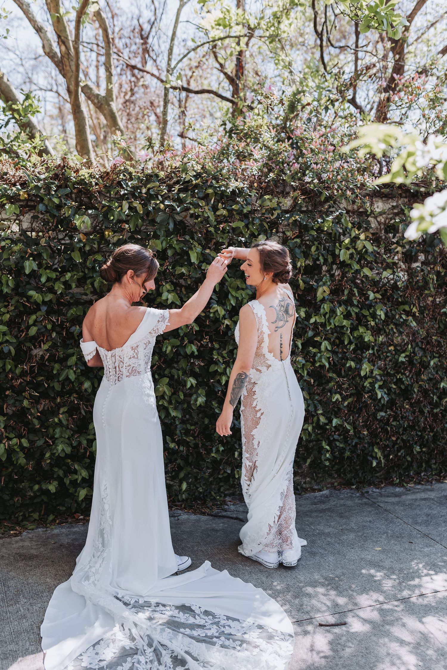 Two brides in white lace wedding gowns dancing together outdoors near an ivy-covered stone wall.
