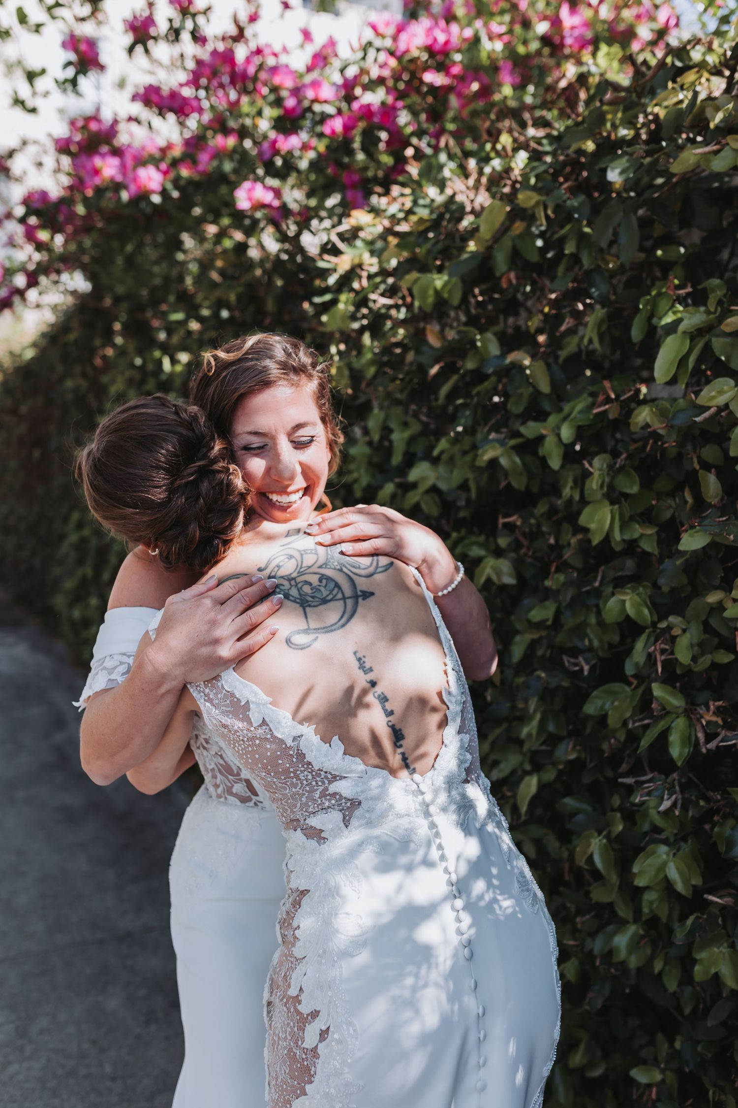 Two brides share a joyful hug in front of vibrant pink flowering bushes on their wedding day.