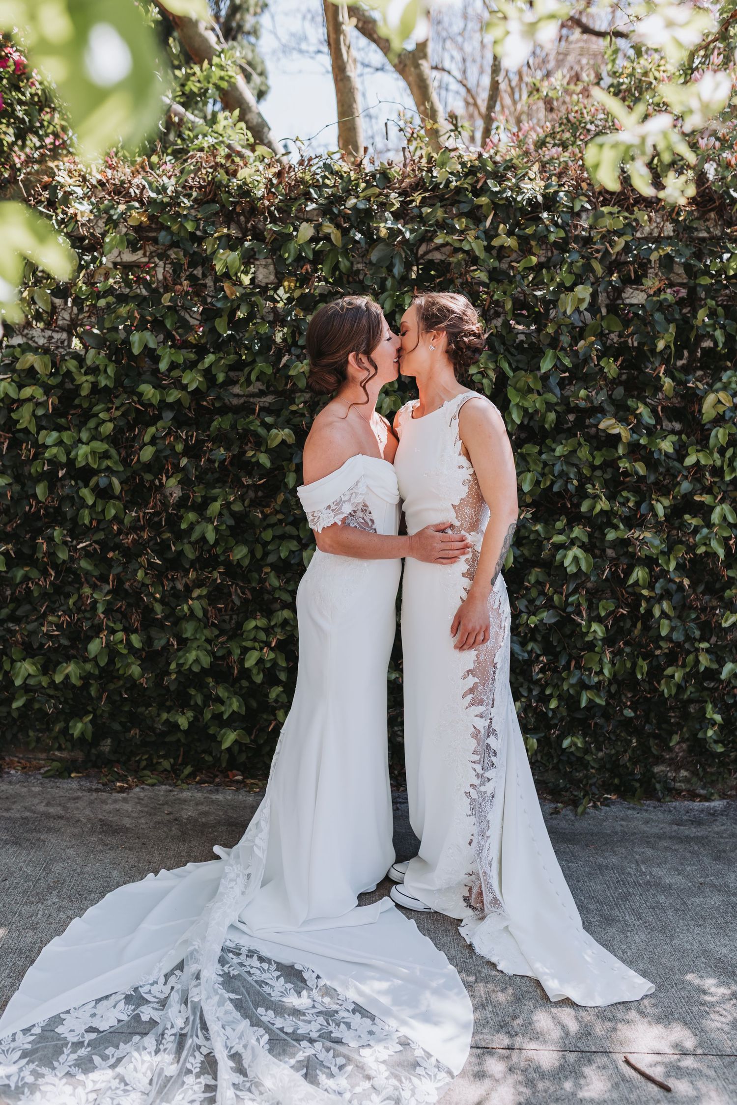 Two brides in elegant white wedding gowns share a kiss in front of a lush green hedge.