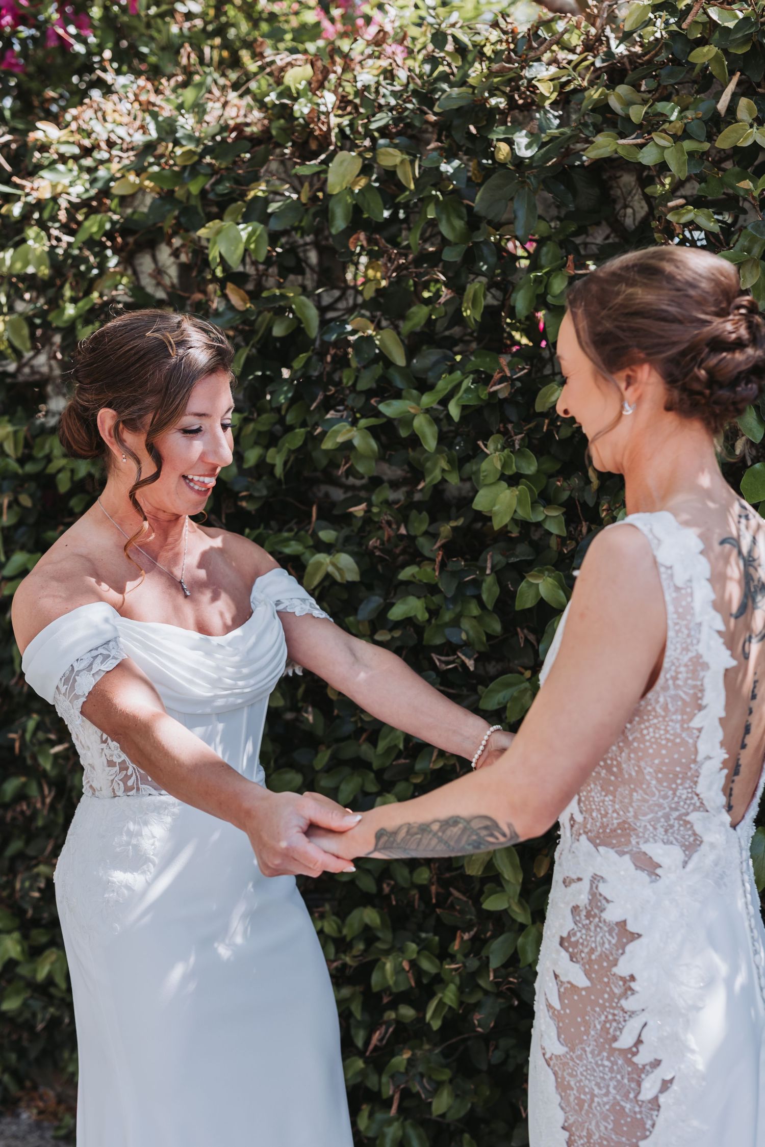 Two brides in white wedding gowns smile and hold hands in front of a lush green garden backdrop.