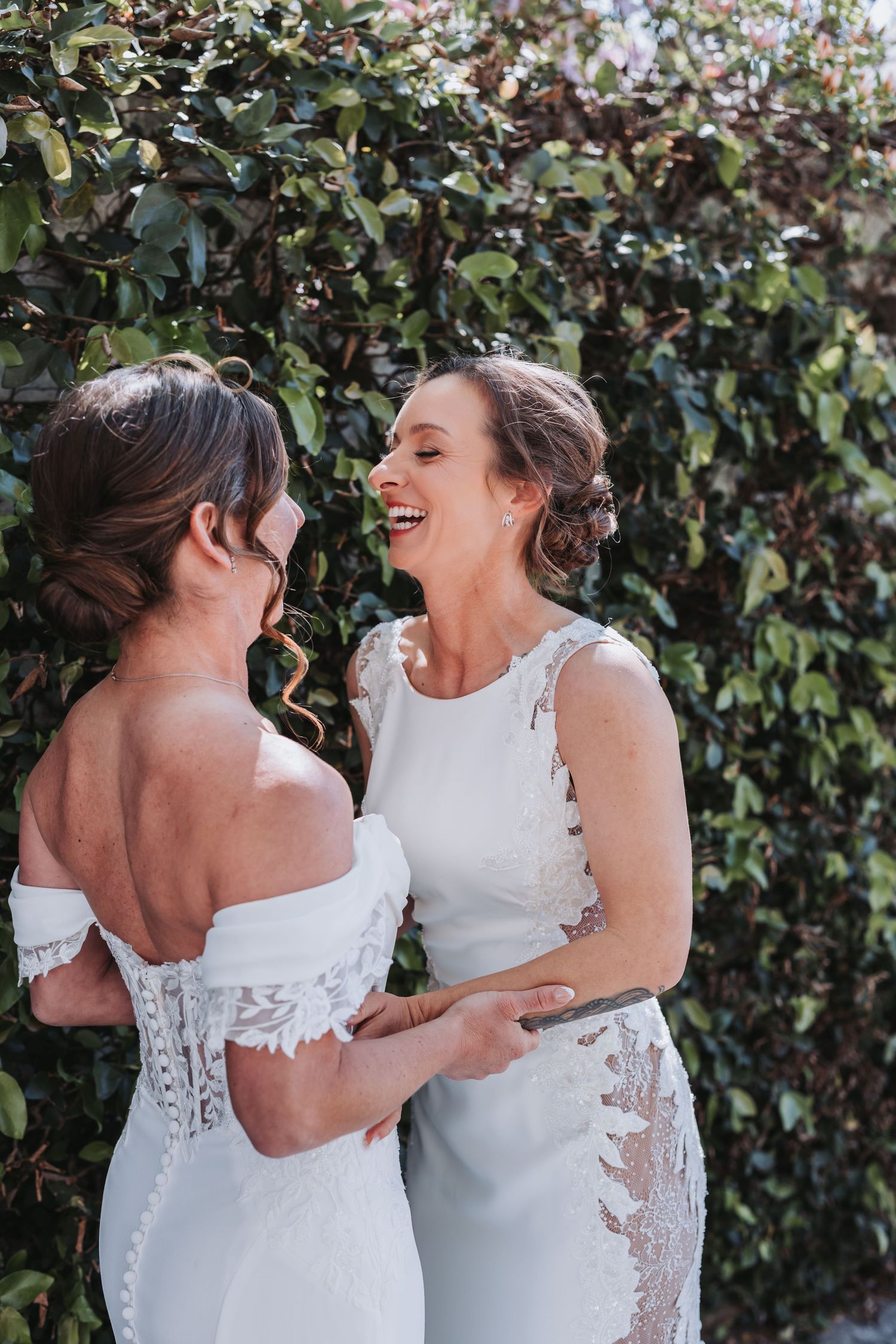 Two brides in white wedding dresses laughing together outdoors against a lush green ivy wall.