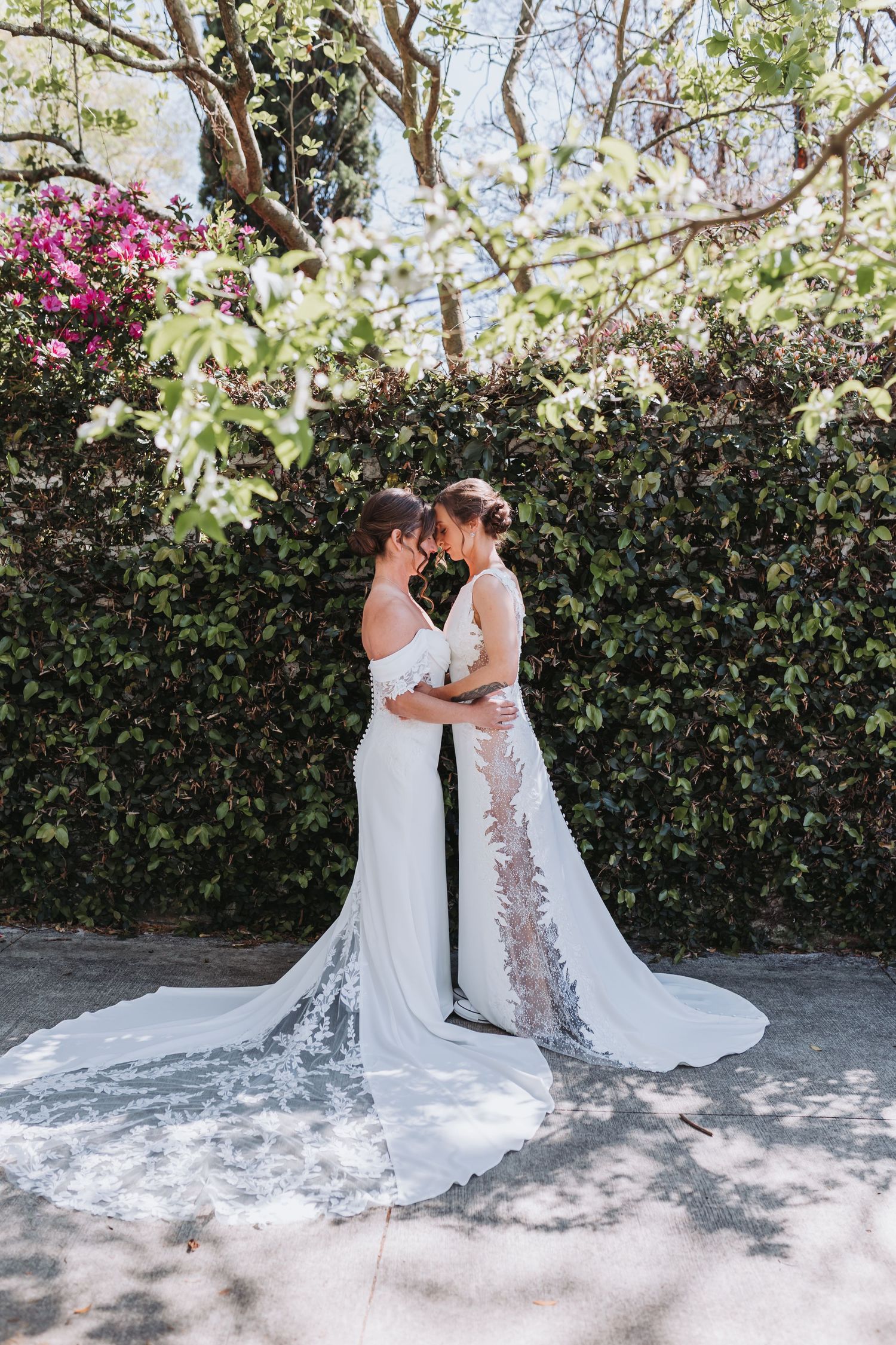 Two brides in white wedding gowns embrace before a lush green ivy wall with pink flowers.