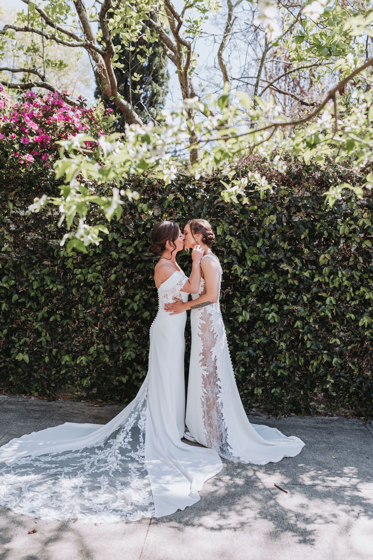 Two brides in white wedding gowns share a kiss in front of a lush ivy wall with pink flowers.