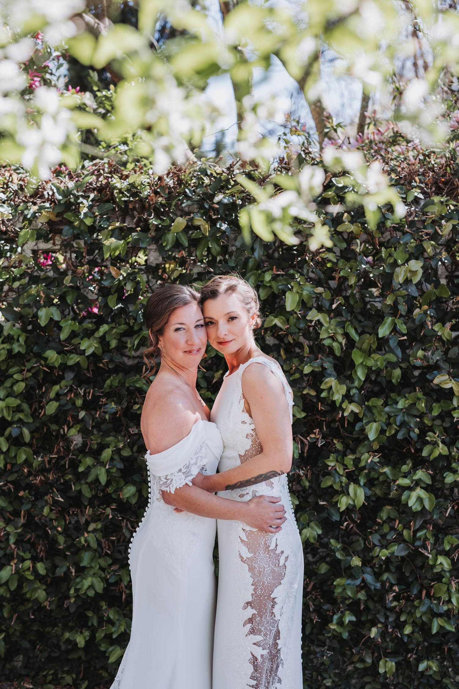 Two brides in elegant white lace wedding dresses hug and smile before a green leafy hedge.