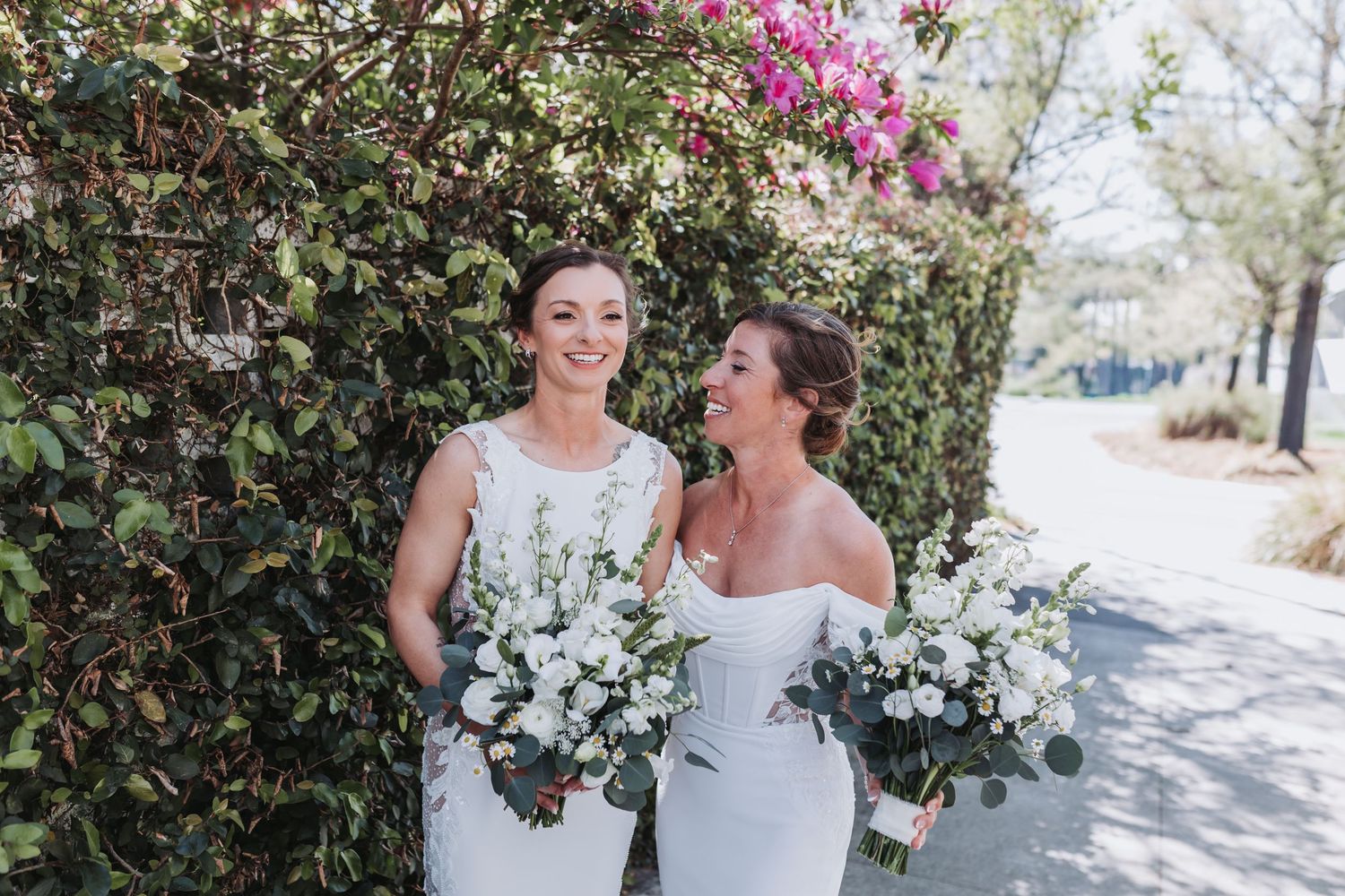 Two brides in white gowns holding bouquets of white flowers, smiling together outdoors near a blooming bougainvillea wall.