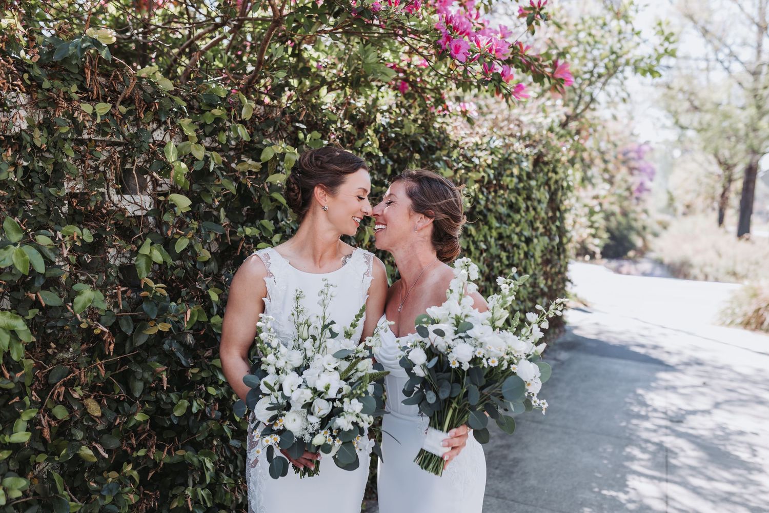 Two brides in white gowns holding white floral bouquets share a tender moment beside a blooming bougainvillea wall.