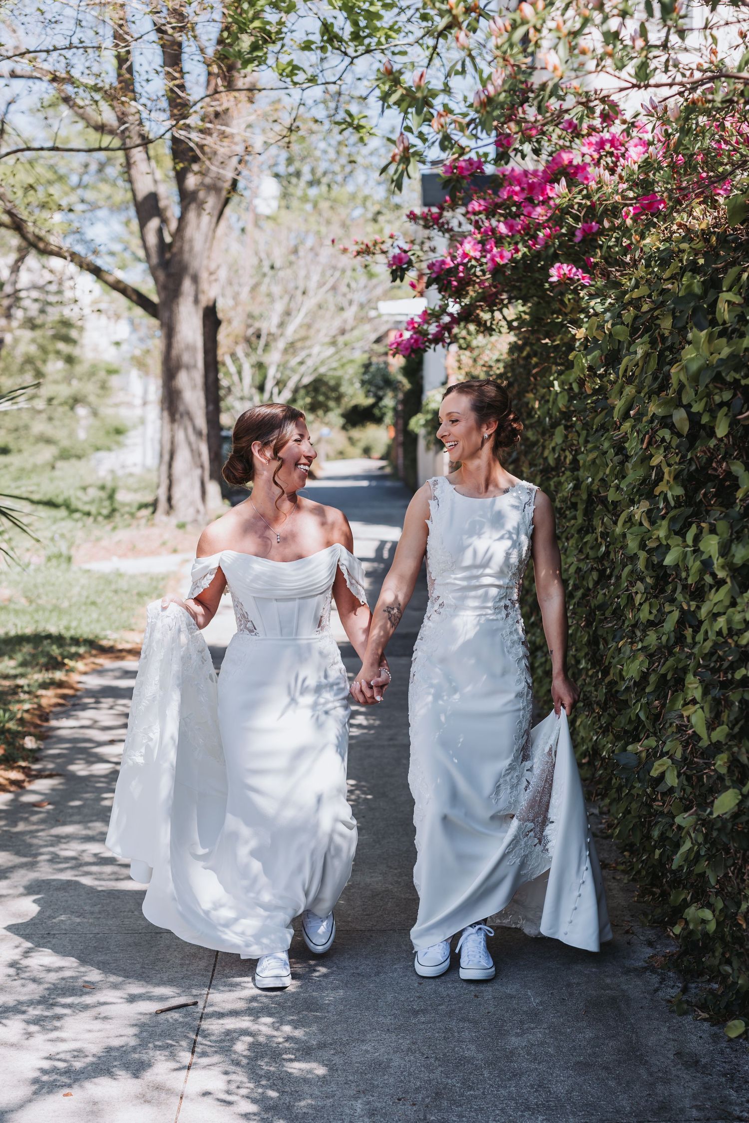 Two brides in white wedding dresses hold hands and walk along a sunlit path lined with pink bougainvillea.