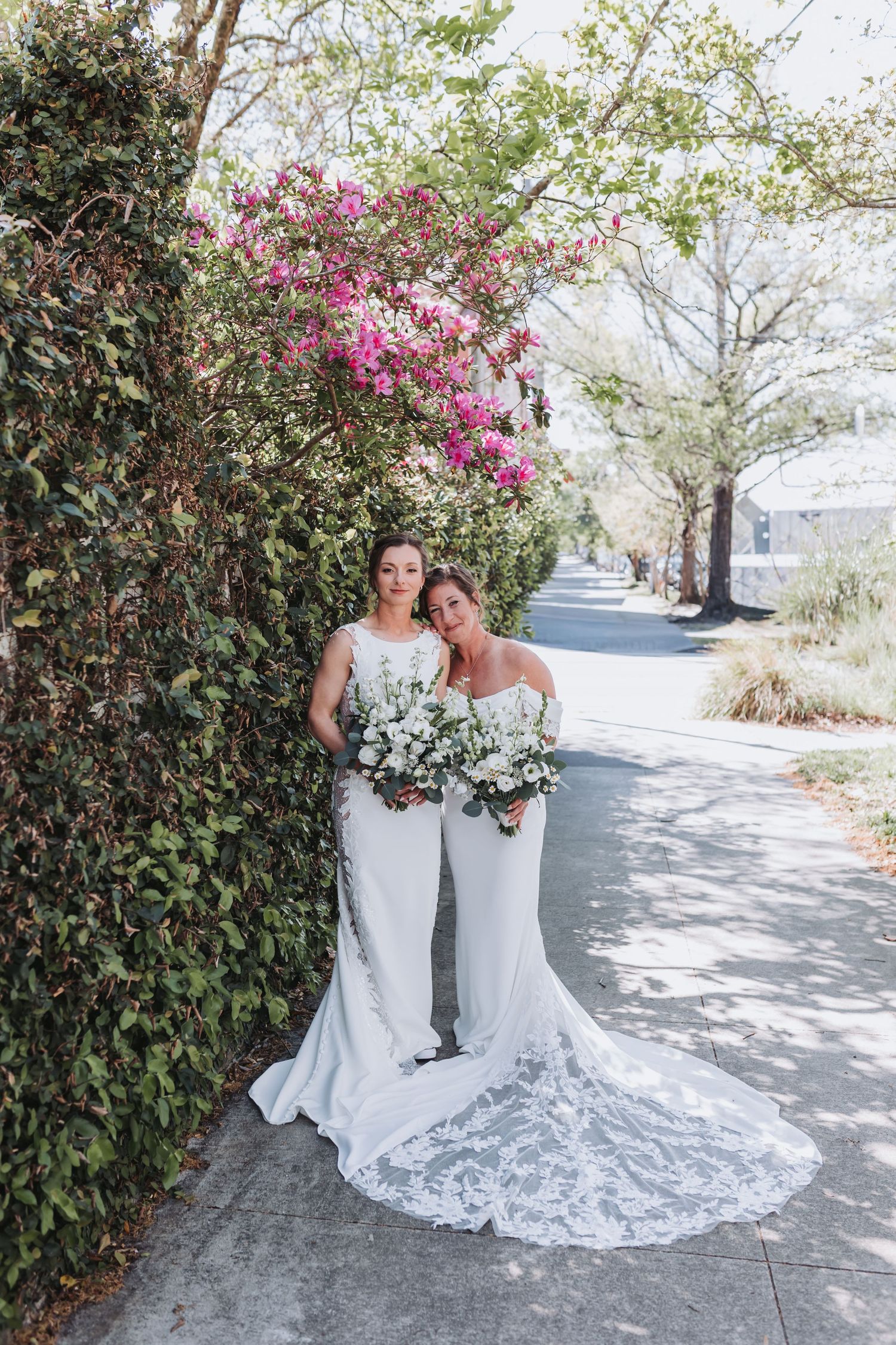 Two brides in white wedding gowns holding bouquets pose on a sidewalk beside a lush ivy wall with pink blooming flowers.