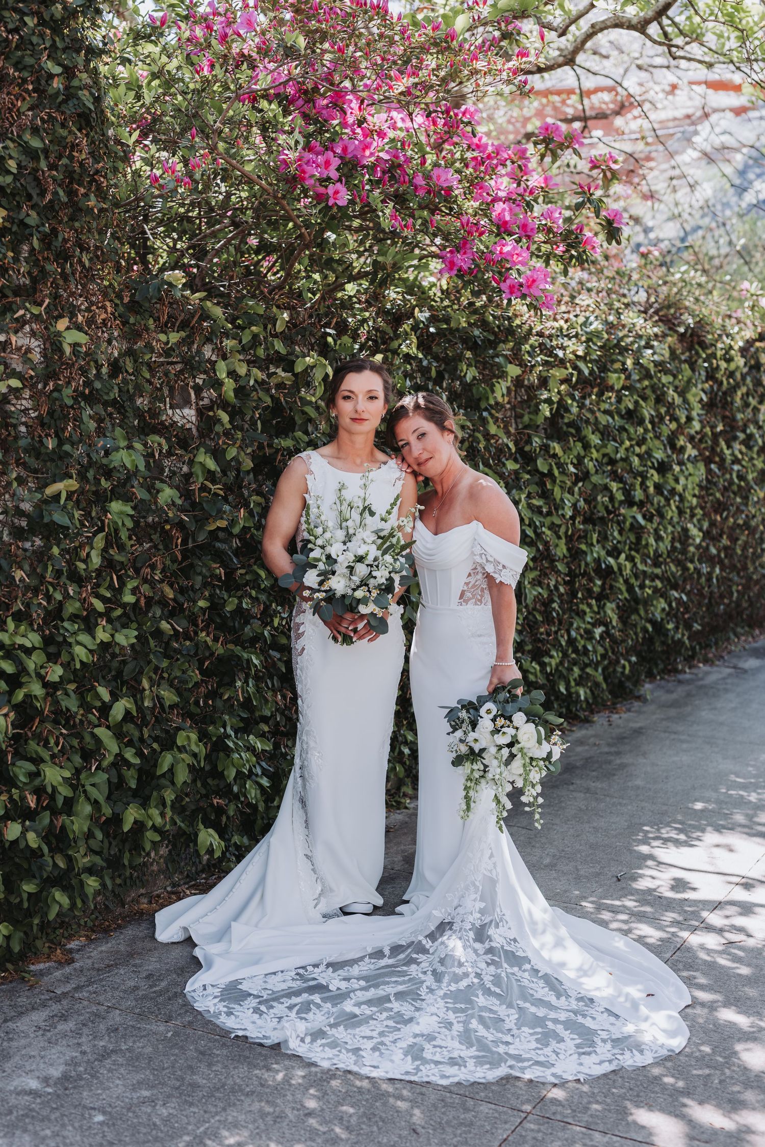 Two brides in white wedding gowns holding bouquets pose before a lush green hedge with pink blooming flowers.