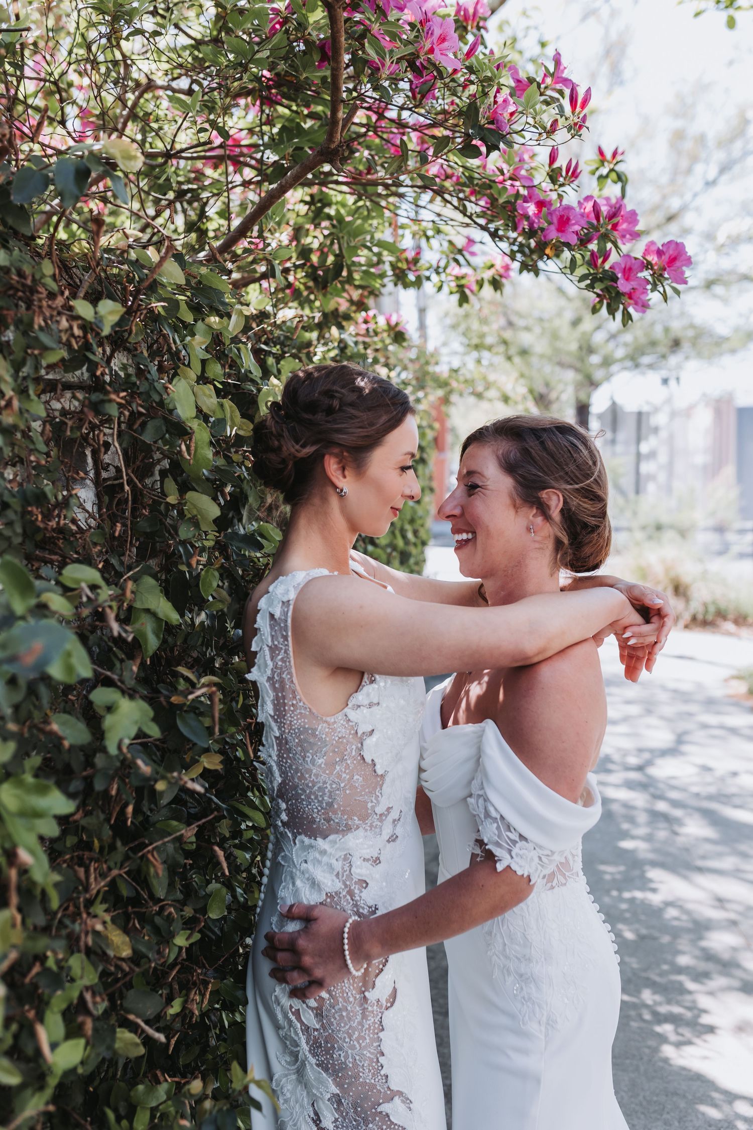 Two brides in white lace wedding gowns embrace and smile under pink flowering branches.