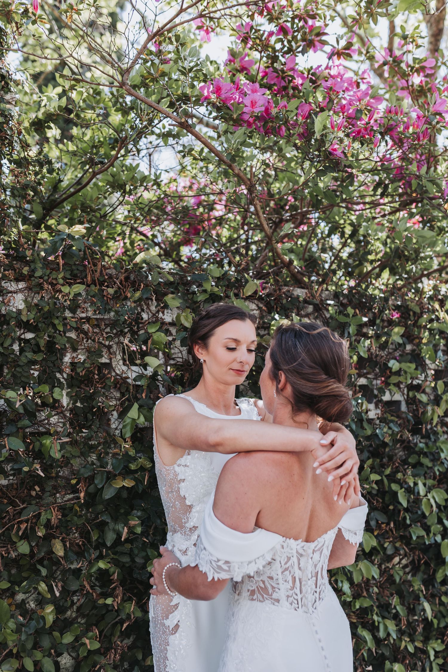 Two brides in white lace wedding gowns embrace tenderly beneath pink blooming flowers outdoors.
