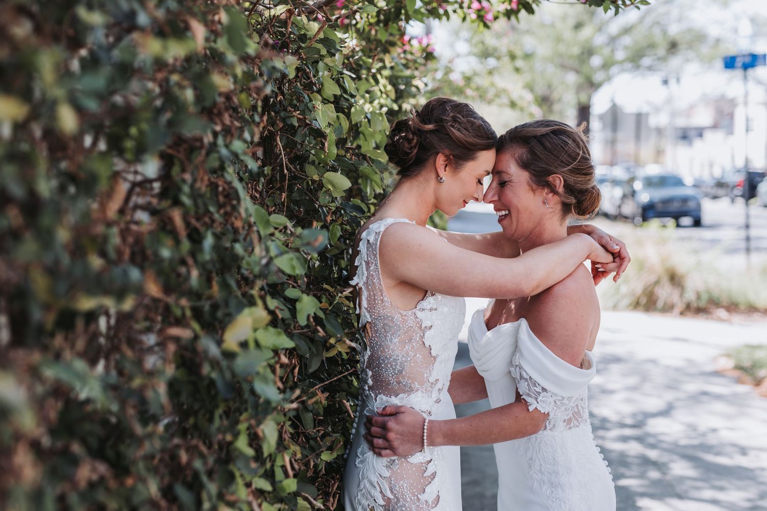 Two brides in white lace gowns embrace tenderly beside an ivy-covered wall outdoors.