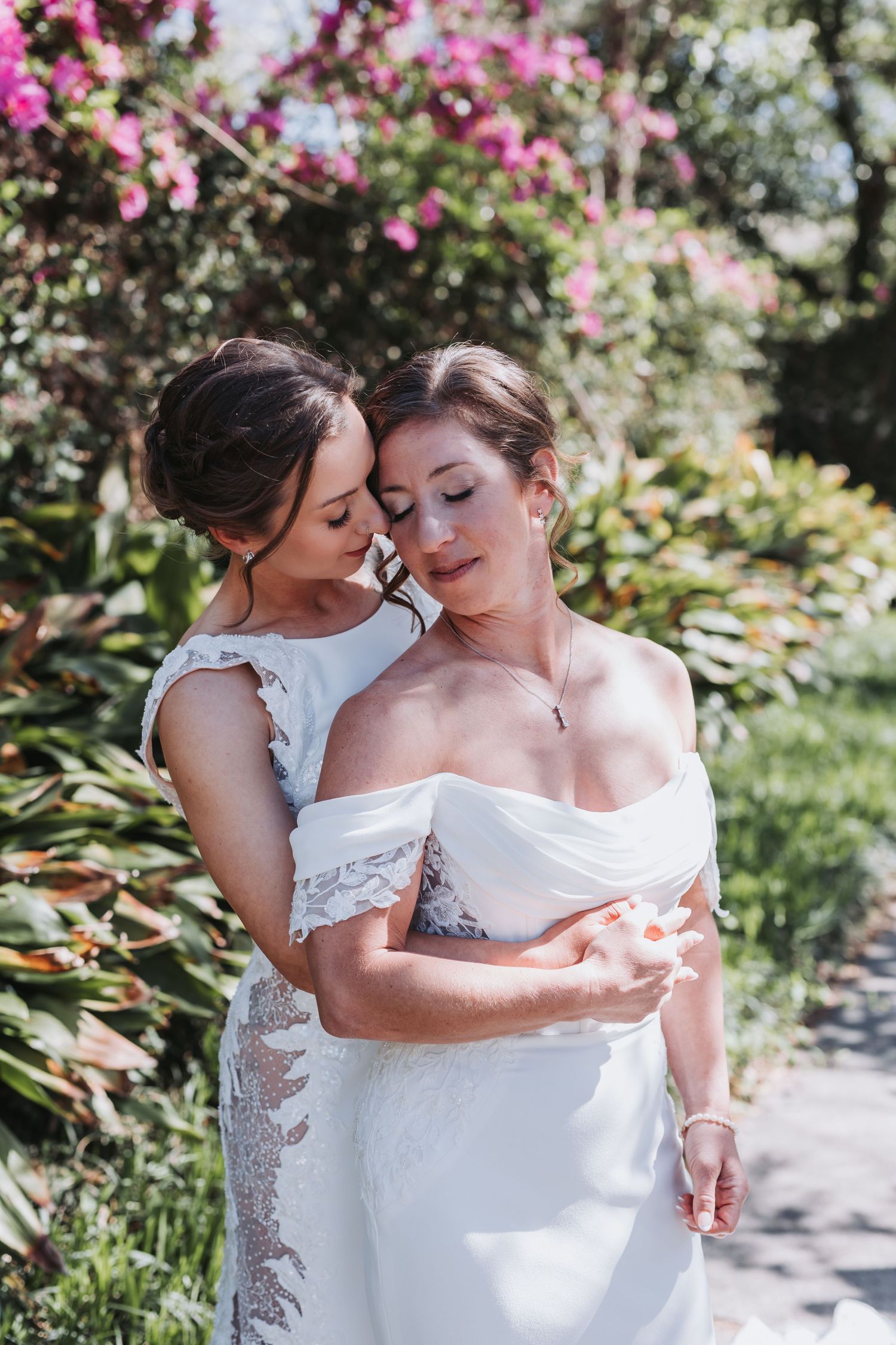 Two brides in white gowns share a tender embrace surrounded by colorful garden flowers.