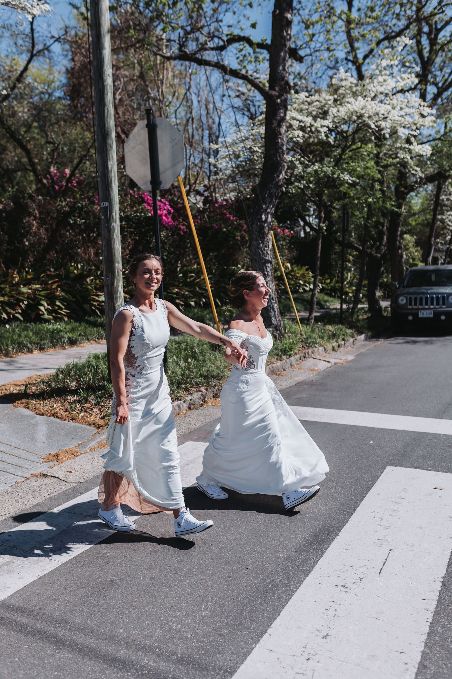 Two brides in white wedding dresses holding hands while crossing a zebra crossing on a sunny tree-lined street.