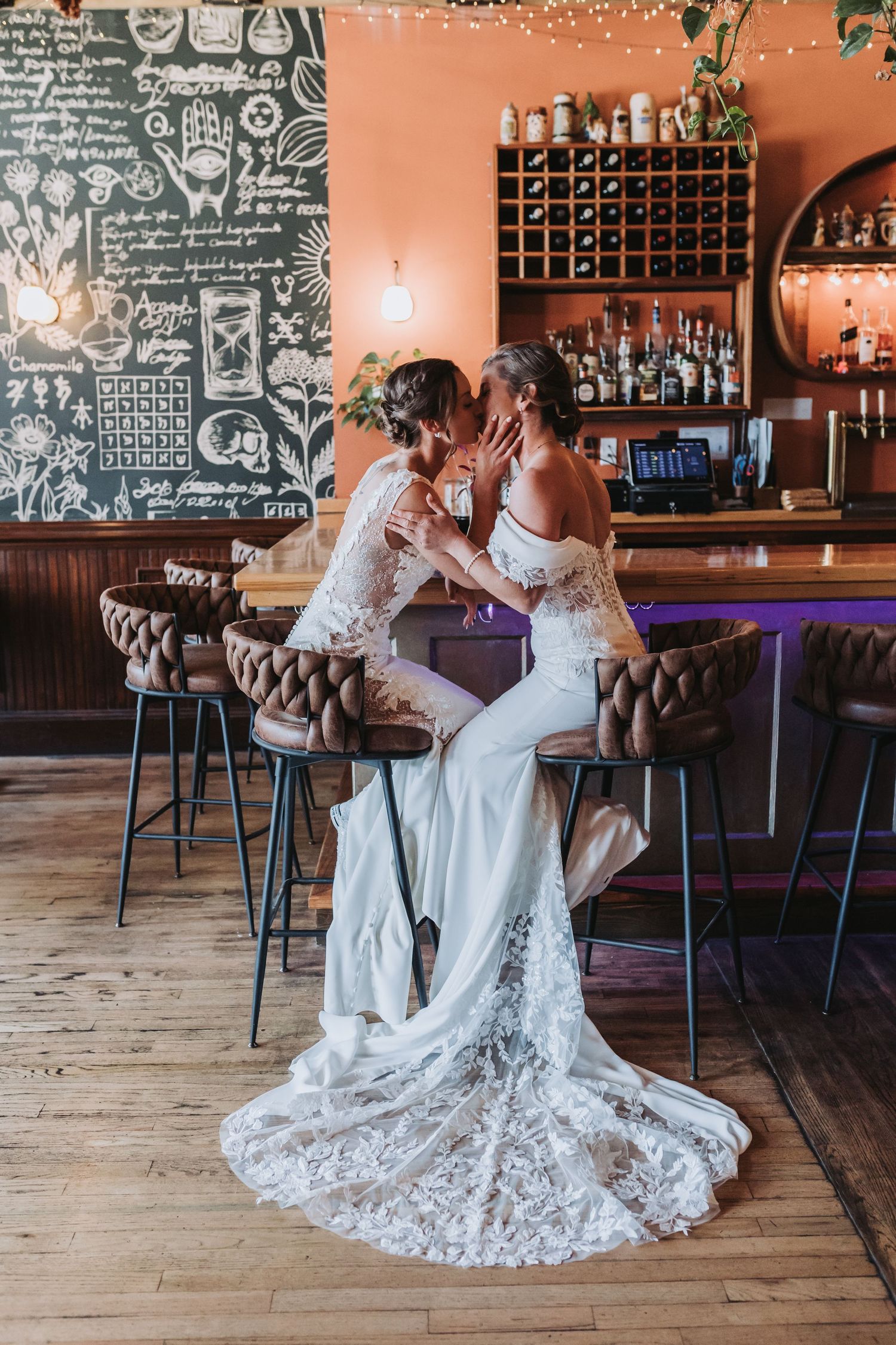 Two brides in white lace wedding dresses share a kiss while seated at a warmly lit bar with a chalkboard wall.