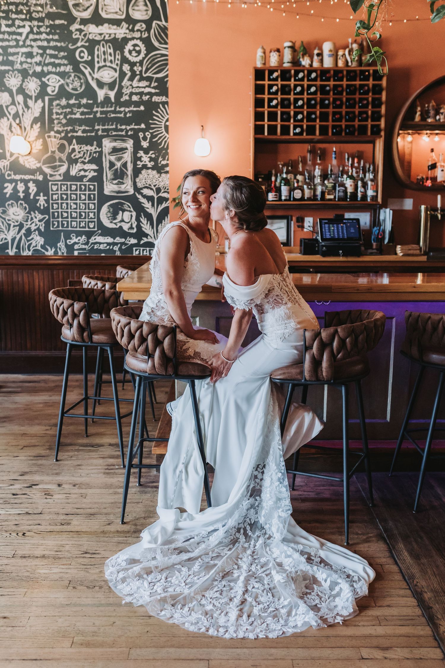 Two brides in wedding gowns share a romantic moment at a stylish bar with chalkboard wall and warm orange decor.