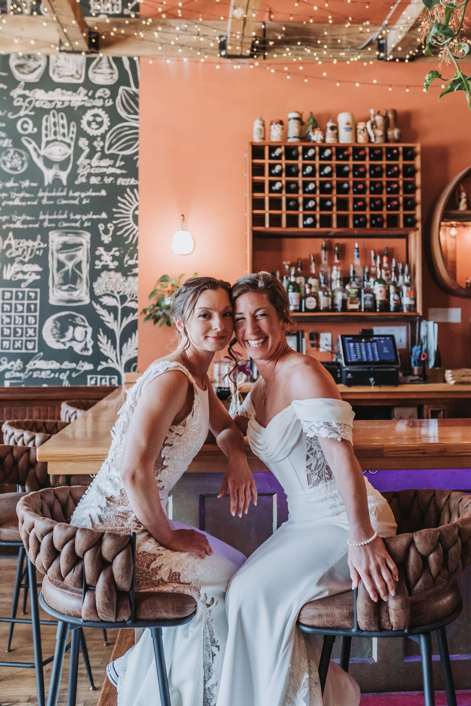 Two brides in white wedding gowns sitting together at a bar, smiling and holding drinks in a warmly lit venue.
