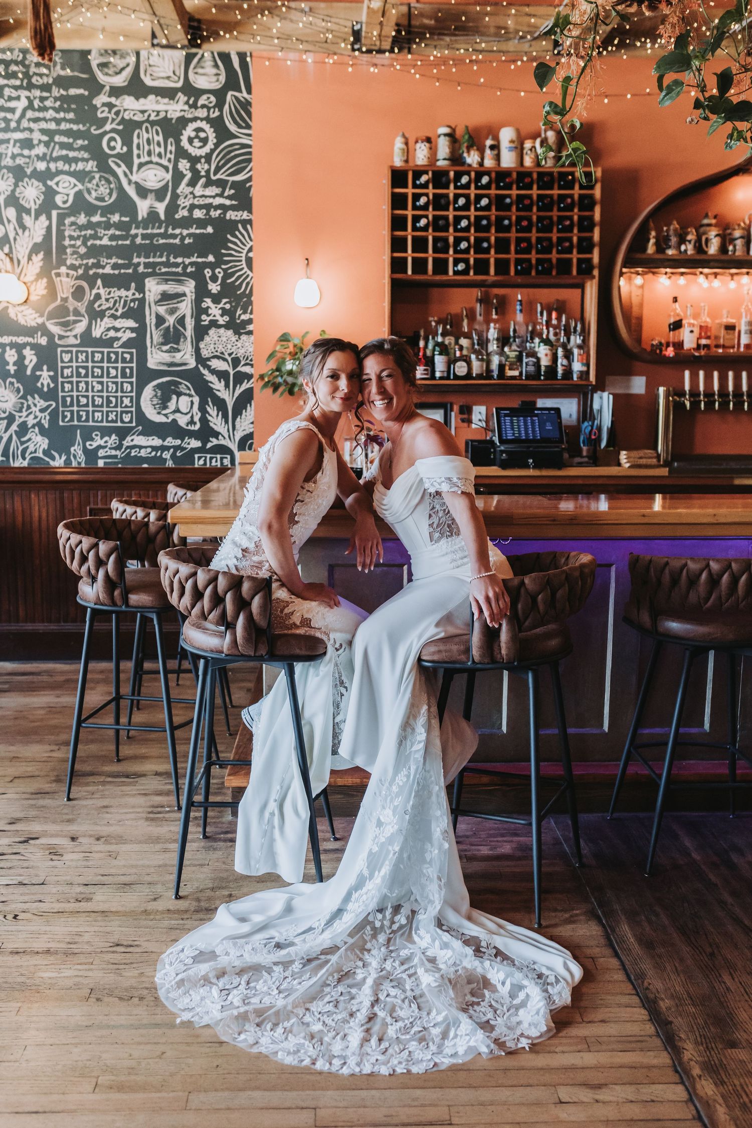 Two brides in white wedding gowns pose together at a warmly lit restaurant bar.