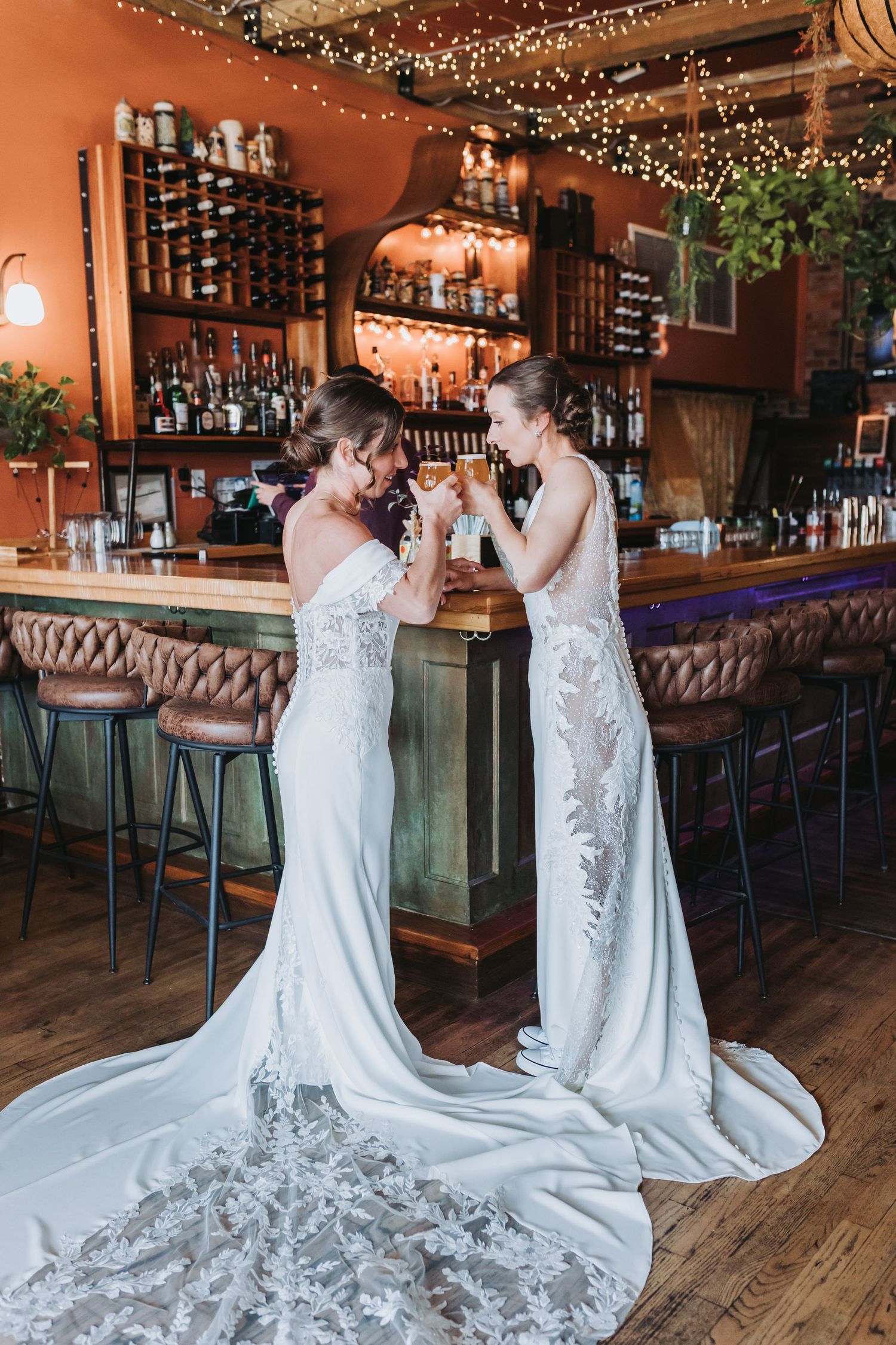 Two brides in elegant white lace wedding gowns toasting drinks at a bar with fairy lights and wine rack backdrop.